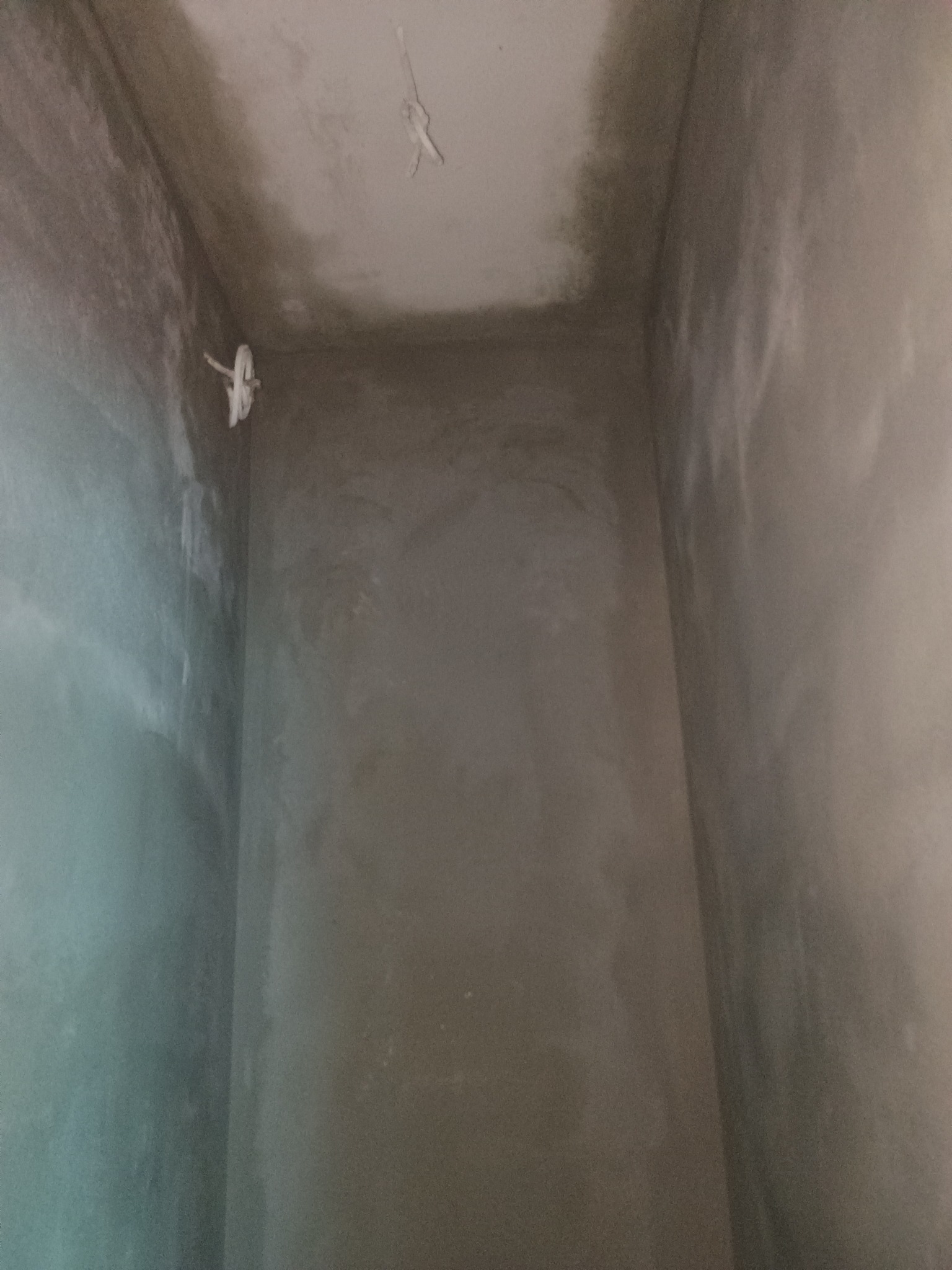 Wnętrze pomieszczenia w trakcie prac wykończeniowych; ściany pokryte świeżym, szarym tynkiem. Widoczne przewody elektryczne wystające ze ściany i sufitu.