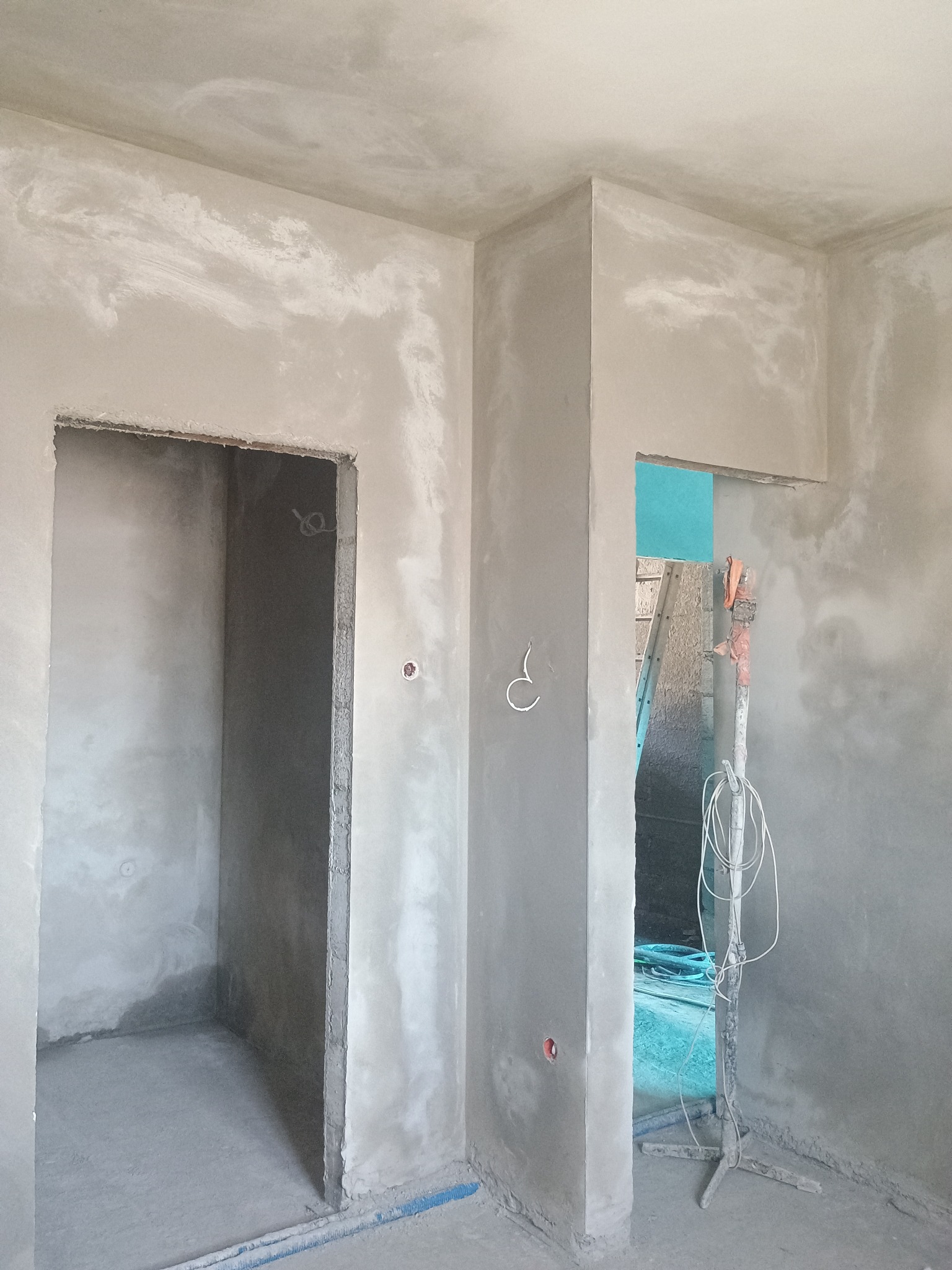 Świeżo nałożona, surowa, szara zaprawa na ścianach i suficie wewnątrz pomieszczenia z widocznymi otworami drzwiowymi, przez jeden z nich widoczna drabina oparta o ścianę.