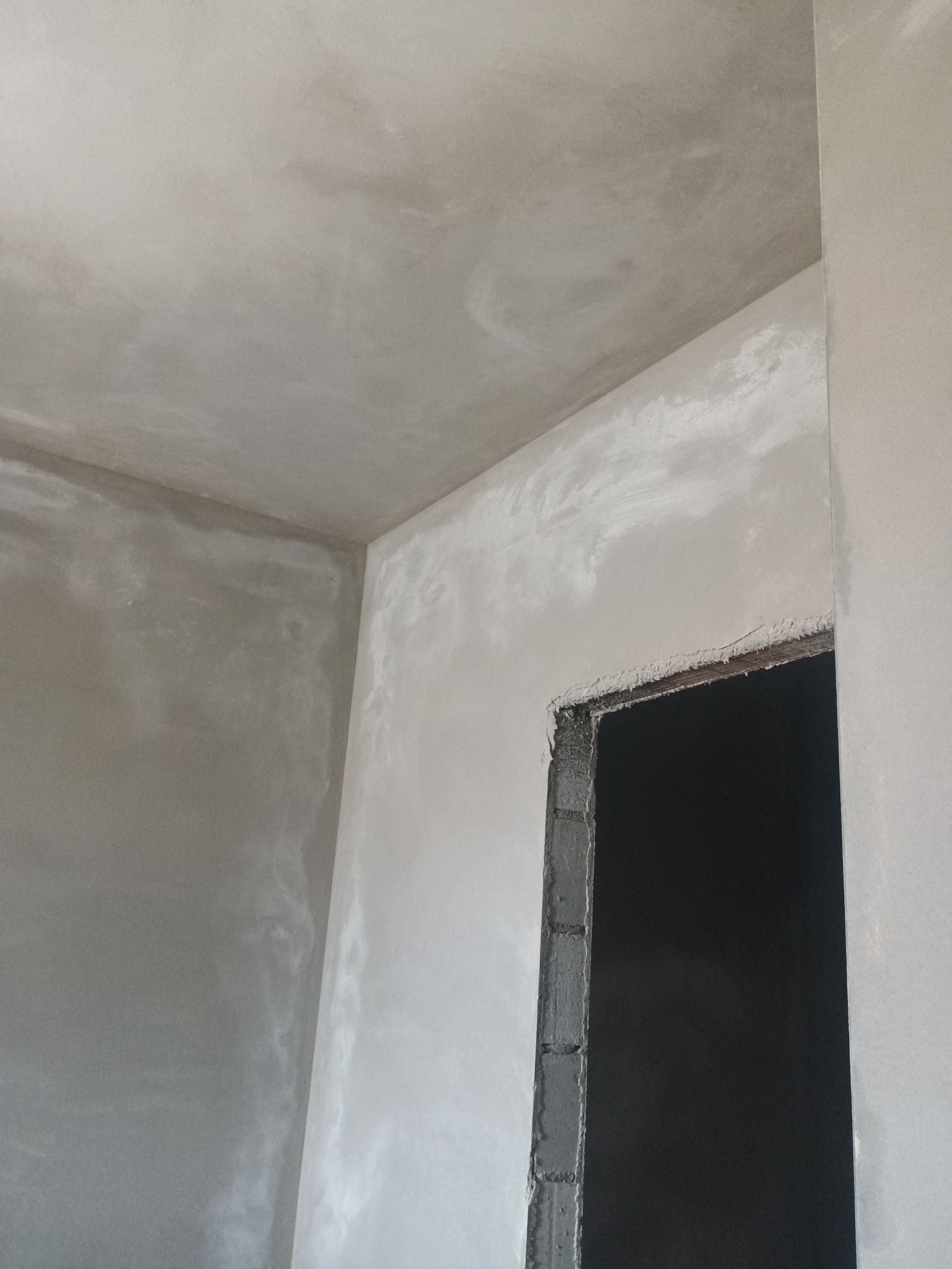 Wnętrze w trakcie prac wykończeniowych, surowe ściany pokryte świeżym, jasnoszarym tynkiem, widoczne nierówności i zacieki, sufit również otynkowany, w tle otwór drzwiowy z surową ramą.