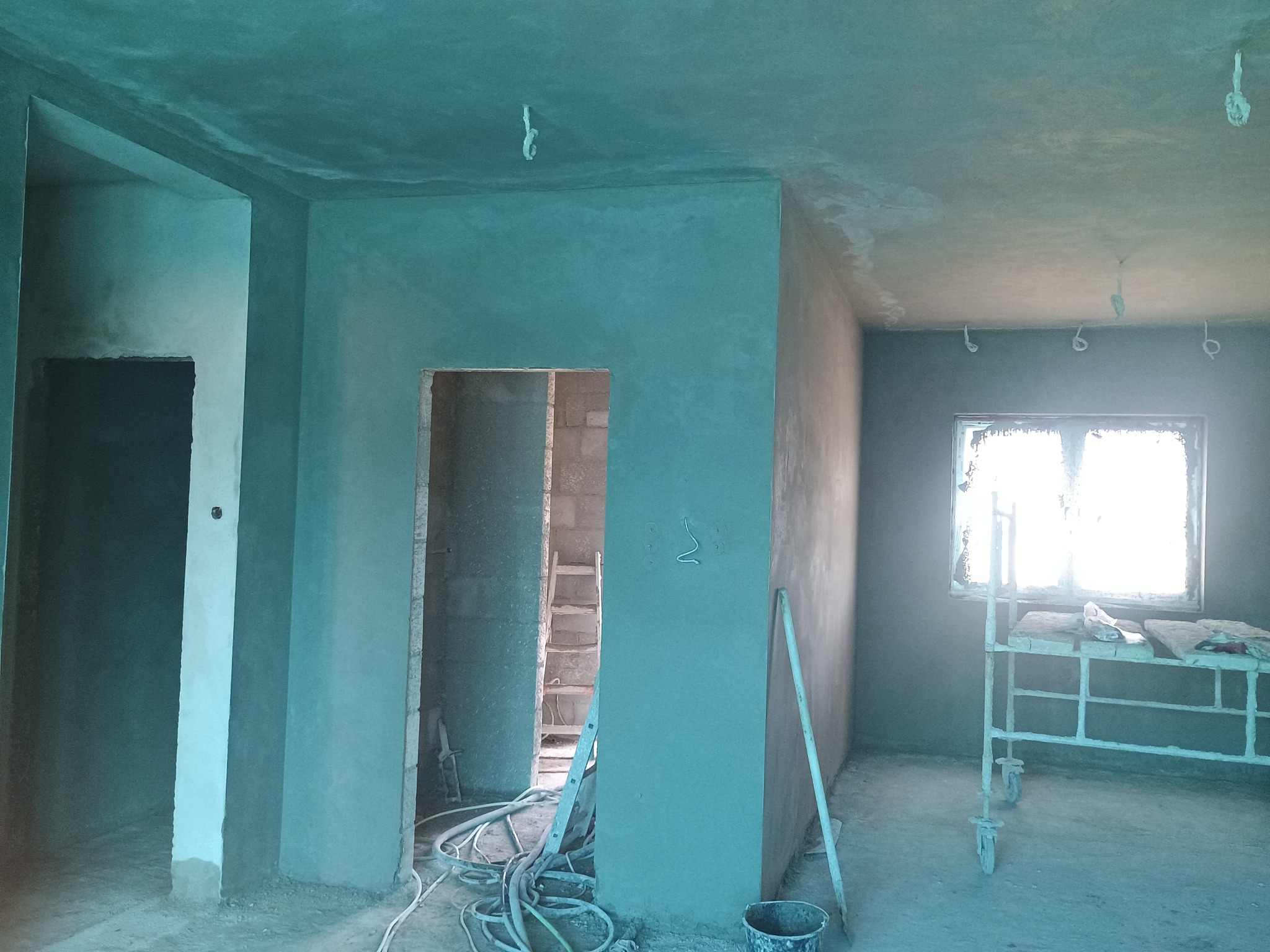 Wnętrze w trakcie prac wykończeniowych: świeżo nałożone tynki w odcieniach szarości i zieleni, widoczne przewody, drabina i rusztowanie pod oknem.