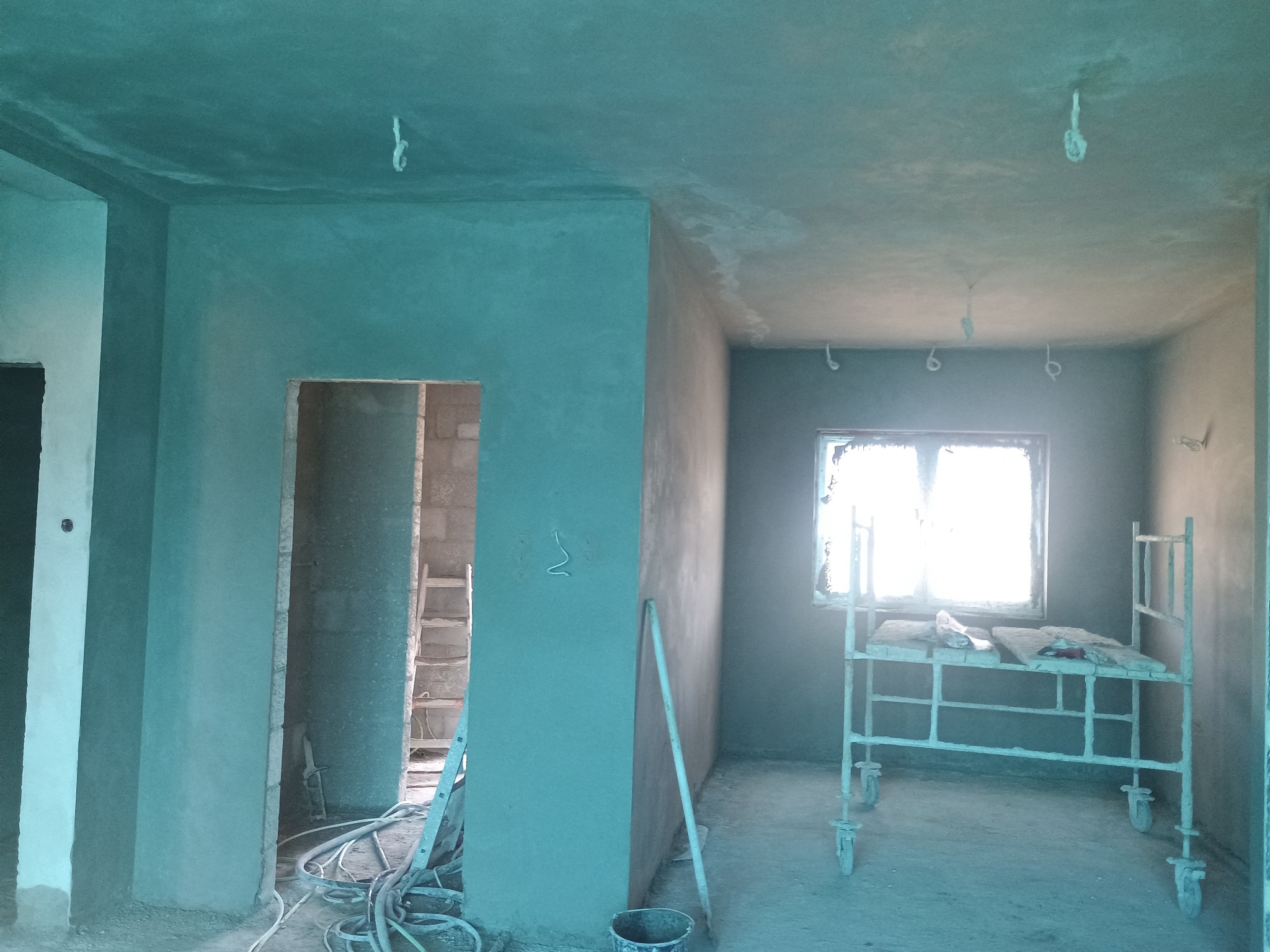 Wnętrze w trakcie prac wykończeniowych, świeżo nałożony tynk na ścianach i suficie, widoczne kable, drabina i rusztowanie na kółkach.