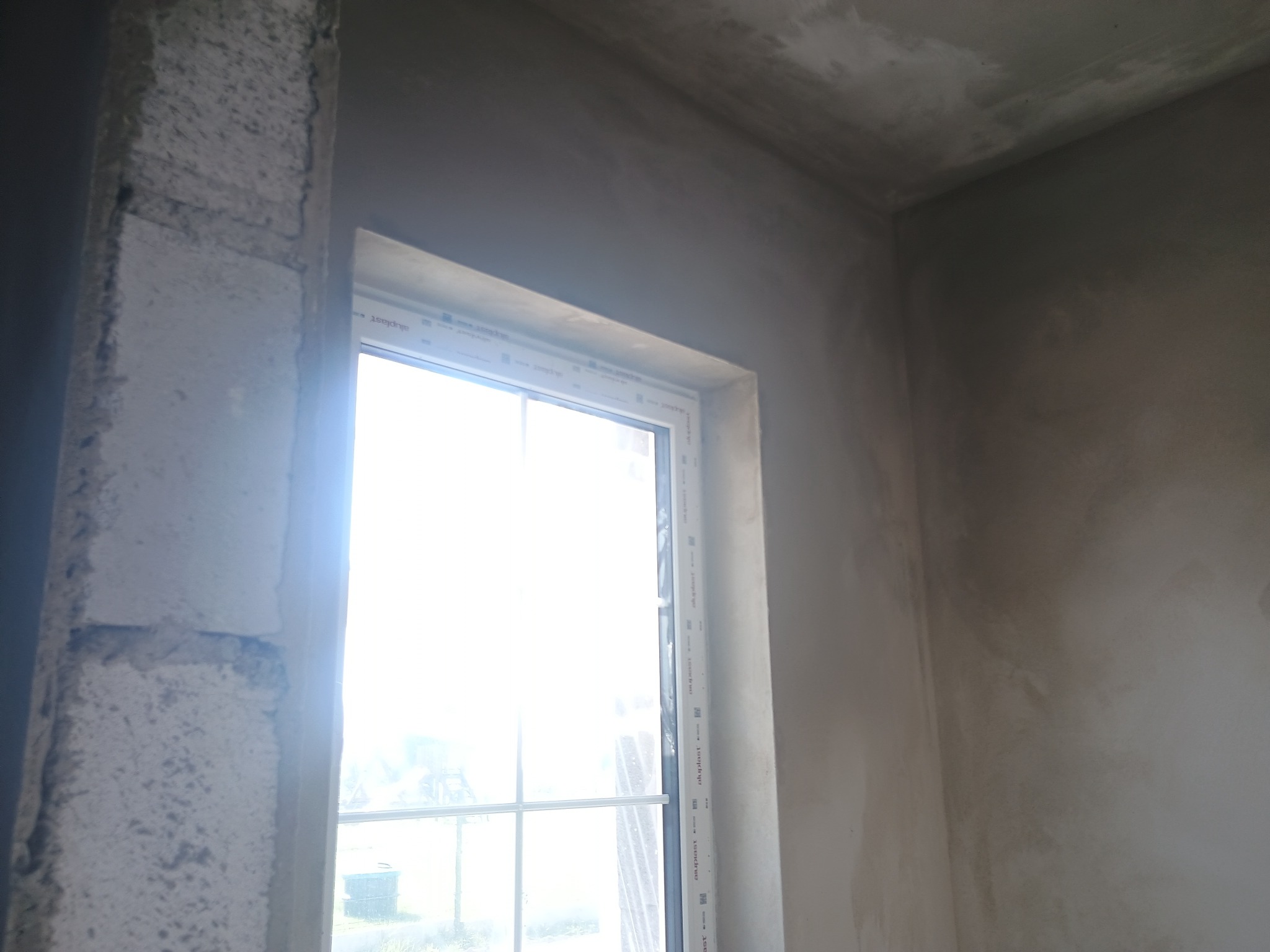 Świeżo otynkowane ściany w narożniku pomieszczenia z oknem, widoczny fragment surowego muru przy ościeżnicy okiennej.