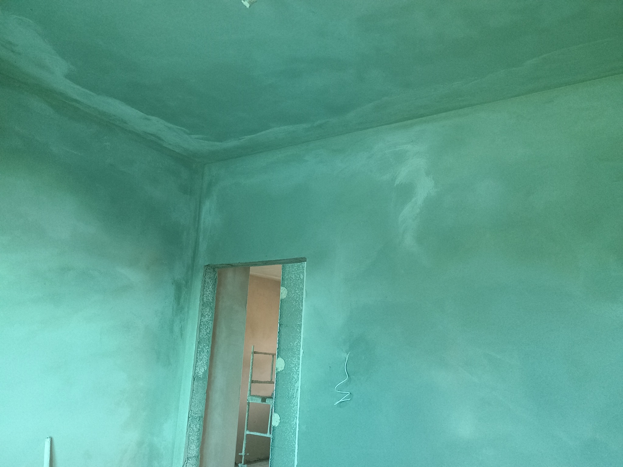 Świeżo nałożona, wilgotna, zielonkawa warstwa tynku na ścianach i suficie, widoczna drabina w przejściu drzwiowym.