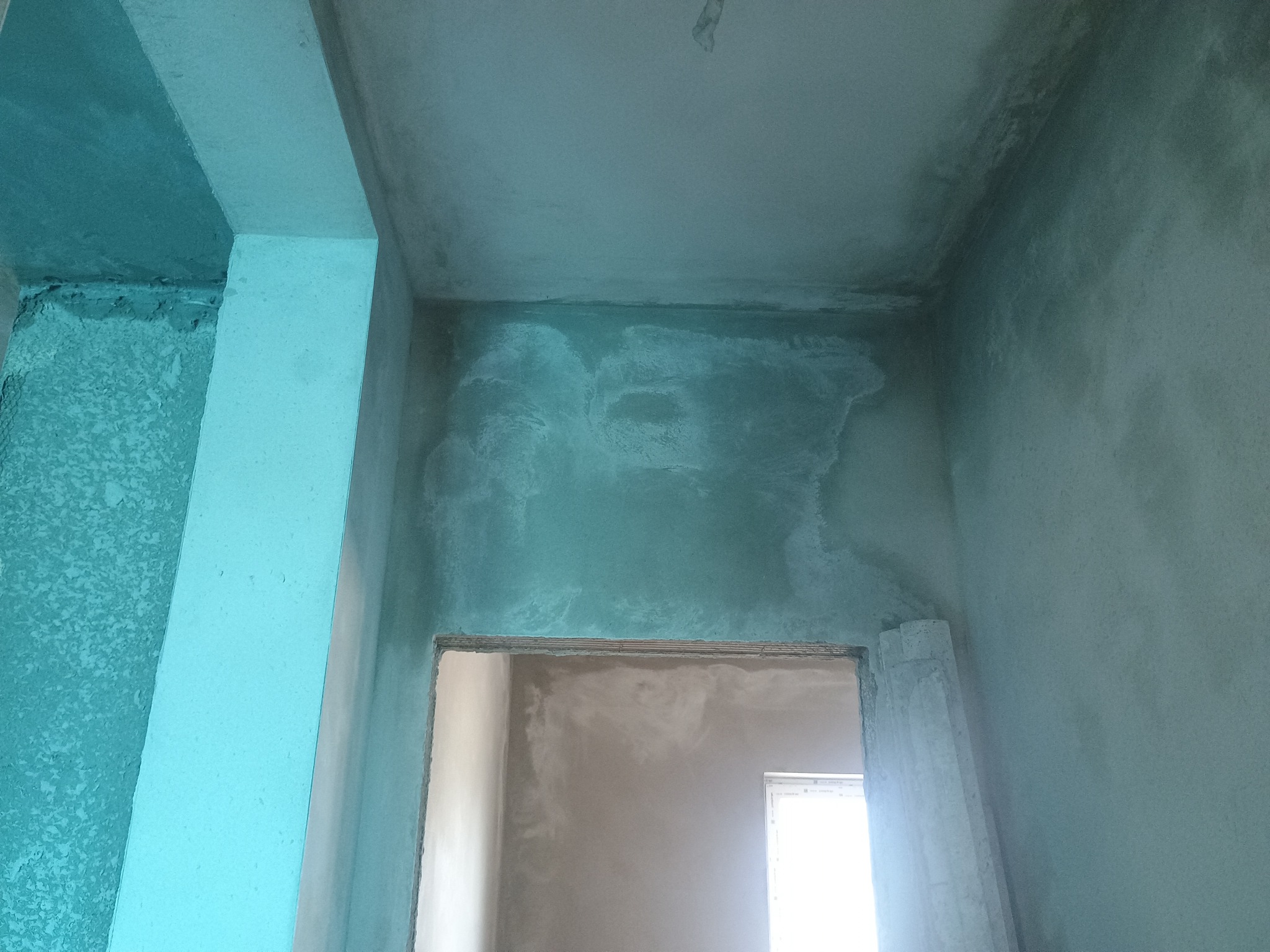 Świeżo nałożona, surowa, szara zaprawa na ścianach i suficie w pomieszczeniu w trakcie remontu, widoczne surowe wykończenie ościeżnicy drzwiowej i fragment okna w tle.