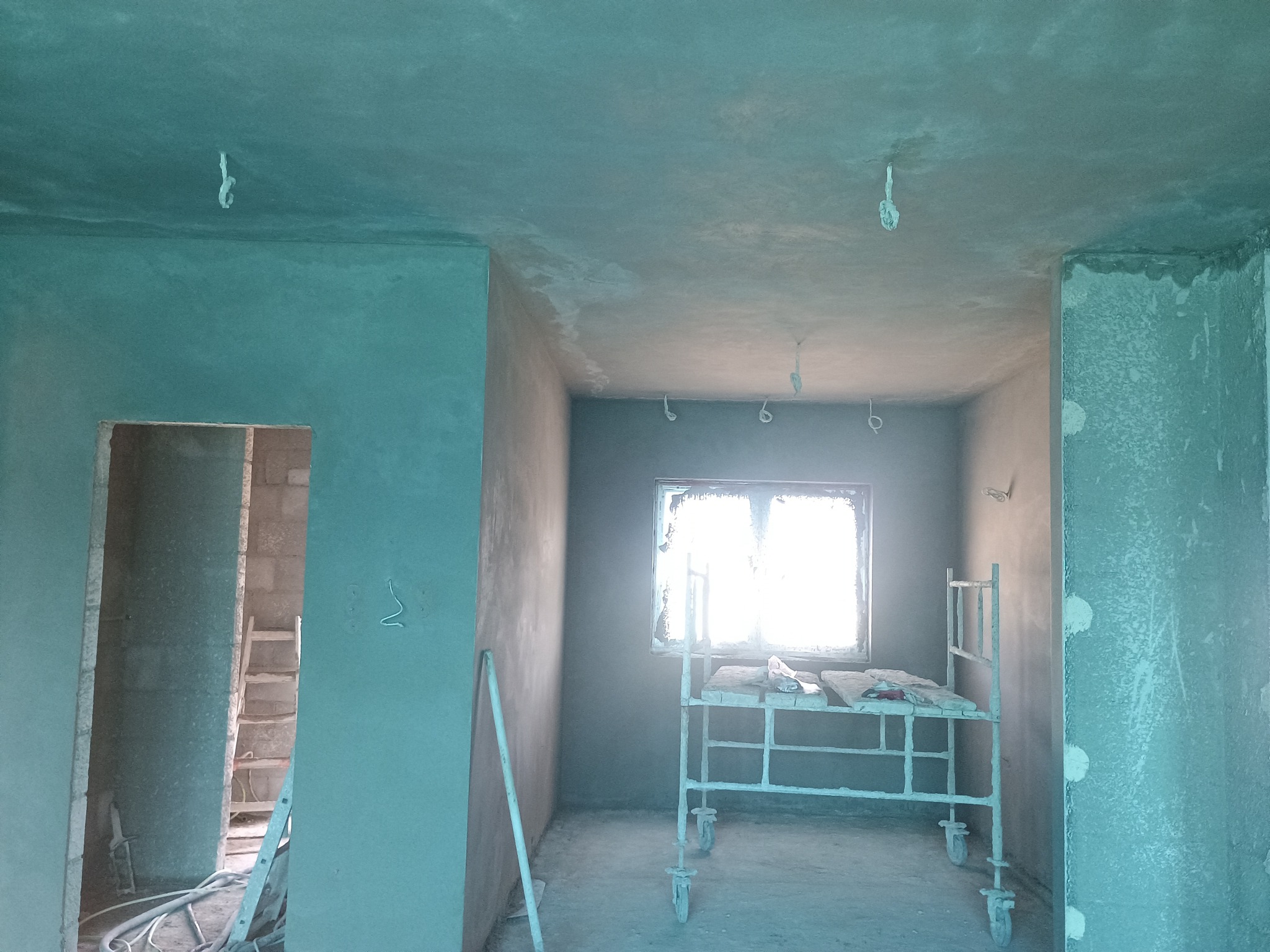Wnętrze w trakcie prac wykończeniowych, świeżo nałożony tynk na ścianach i suficie, widoczne rusztowanie przy oknie i drabina w przejściu do innego pomieszczenia.