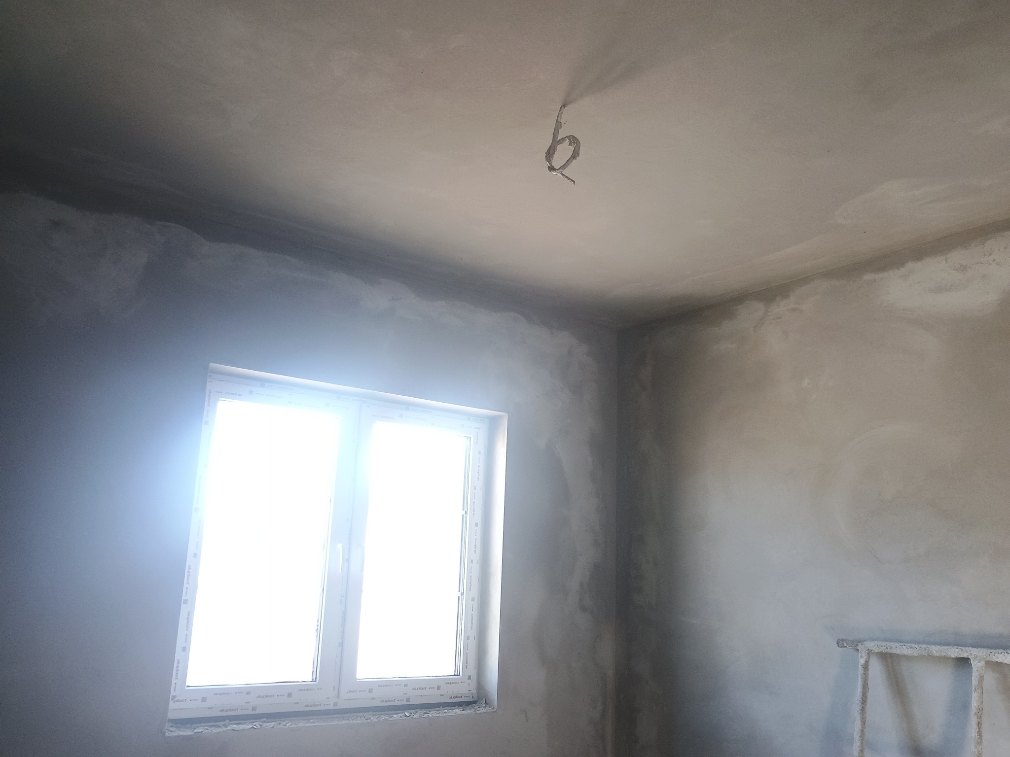 Świeżo otynkowane, surowe wnętrze z przewodem elektrycznym zwisającym z sufitu i jasnym oknem po lewej stronie.