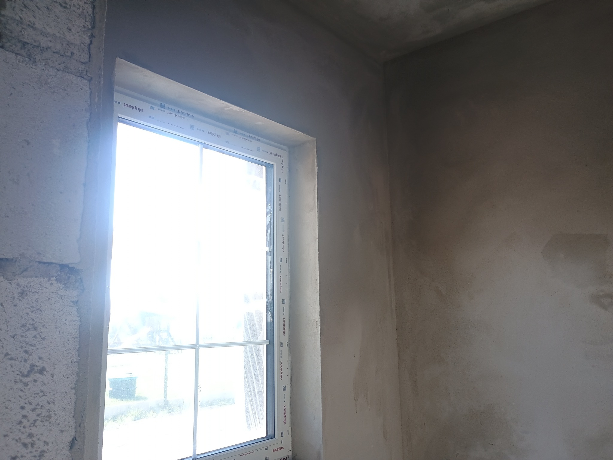 Surowe, szare ściany wewnętrzne w trakcie prac wykończeniowych, widoczne ślady po tynkowaniu przy oknie z białymi ramami.