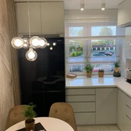 Nowoczesna, jasna kuchnia z białymi, błyszczącymi szafkami, okrągłym stołem z beżowymi krzesłami i dekoracyjnym oświetleniem sufitowym w postaci lampy z kulistymi żarówkami. Na blacie pod oknem...