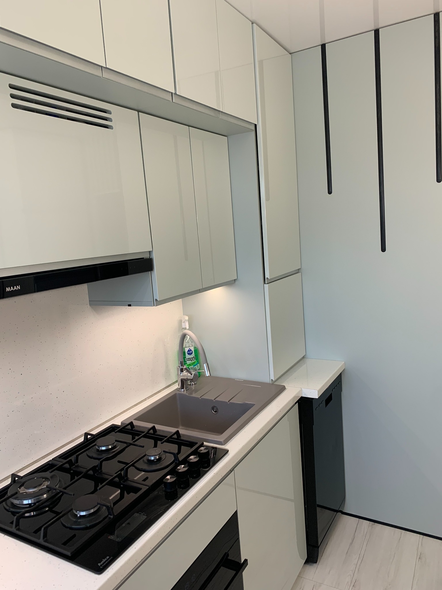 Nowoczesna kuchnia z białymi, lakierowanymi frontami, czarną płytą grzewczą i zlewozmywakiem w odcieniach szarości, uzupełniona o czarne, minimalistyczne uchwyty i sprzęt AGD.