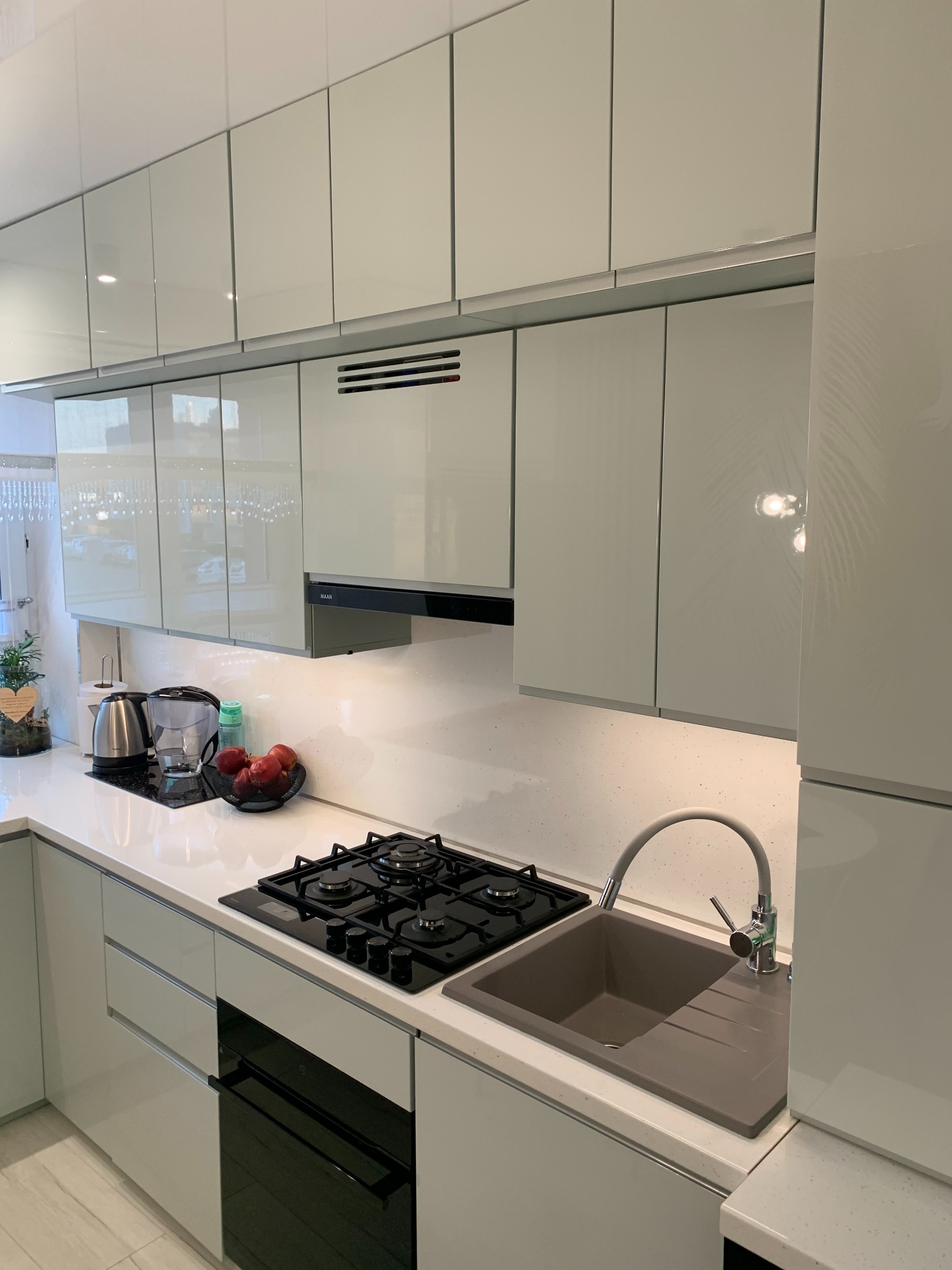 Nowoczesna kuchnia z białymi, lakierowanymi frontami szafek, czarną płytą grzewczą i szarą, kompozytową umywalką z baterią w odcieniach szarości, uzupełniona o czajnik elektryczny, dzbanek...