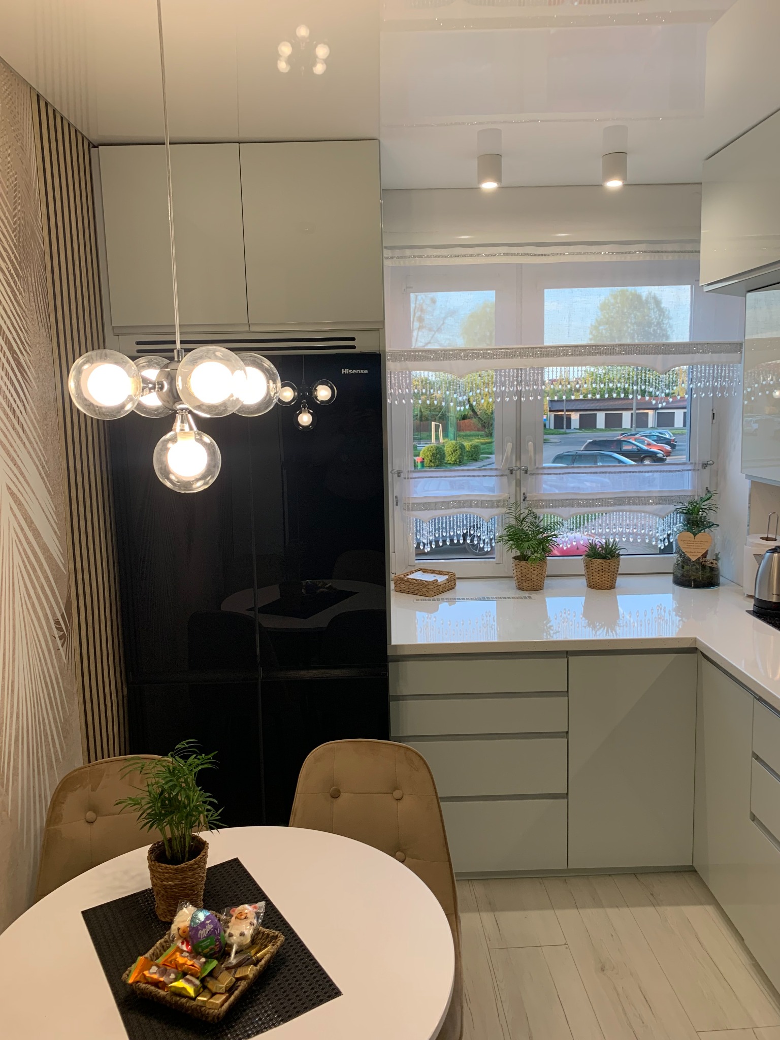 Nowoczesna, jasna kuchnia z białymi, błyszczącymi szafkami, okrągłym stołem z beżowymi krzesłami i dekoracyjnym oświetleniem sufitowym w postaci lampy z kulistymi żarówkami. Na blacie pod oknem...