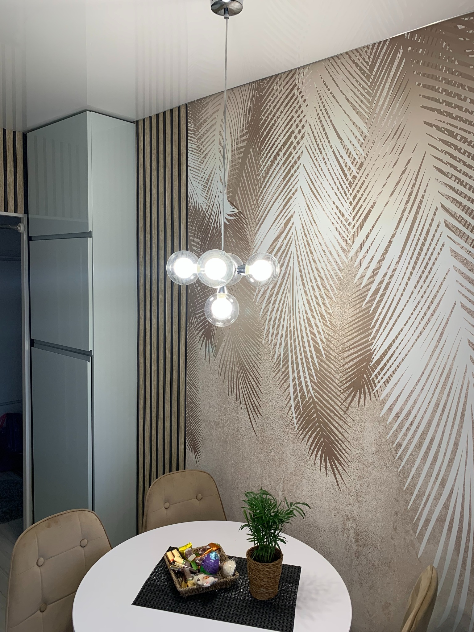 Jadalnia z okrągłym białym stołem, beżowymi krzesłami, lampą sufitową z pięcioma szklanymi kulami i ścianą z motywem palmowych liści, widoczny fragment szafy.