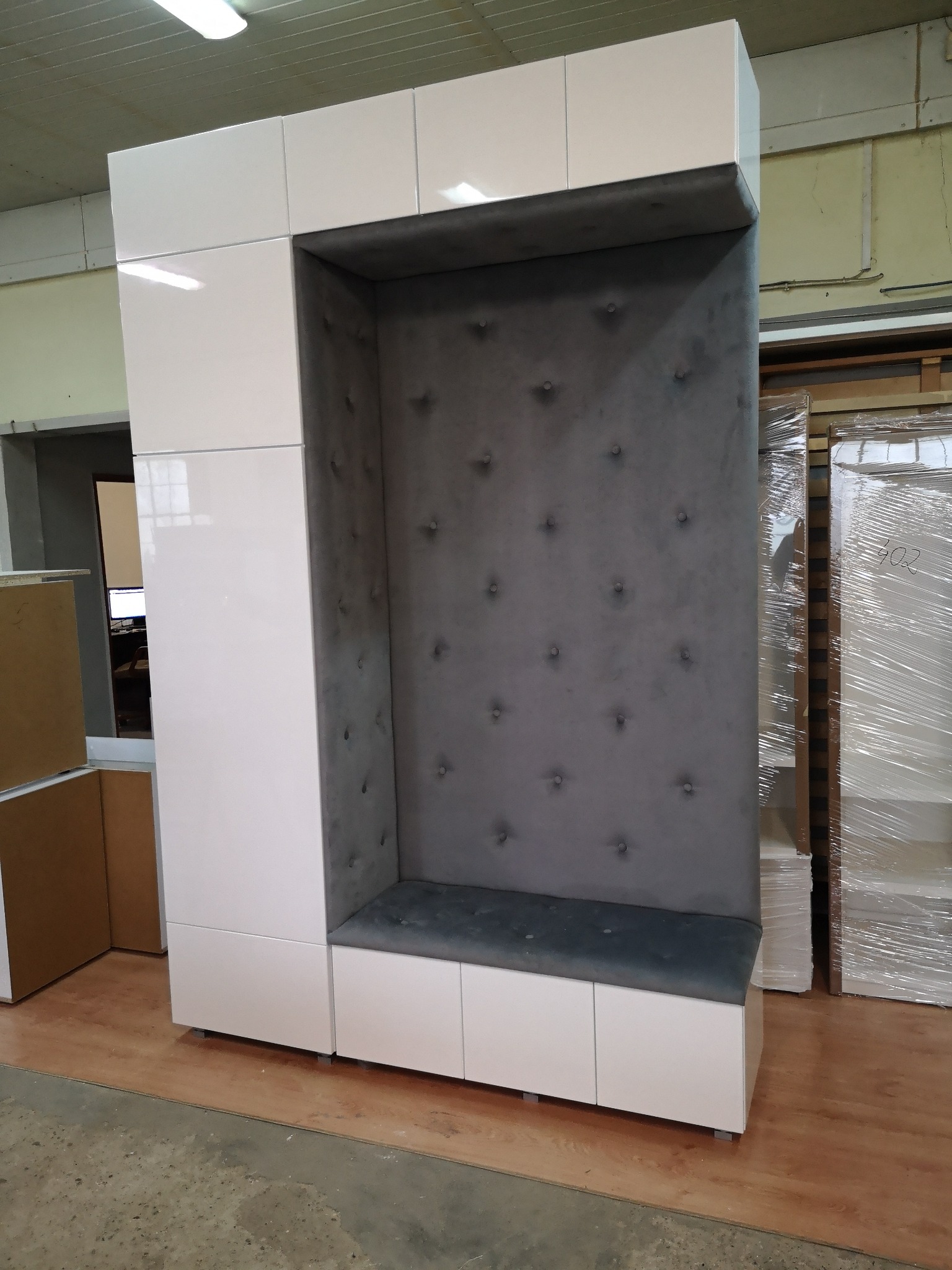 Biała szafa z wnęką wyściełaną szarym materiałem z guzikami, połączona z siedziskiem i szafkami dolnymi.