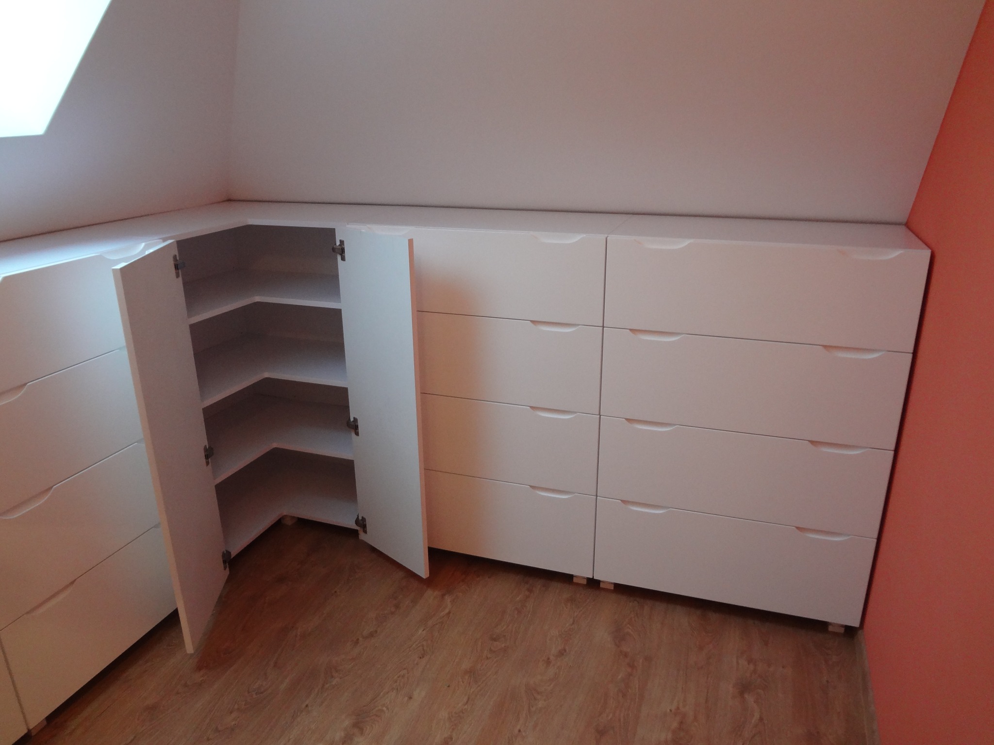 Biała szafa wnękowa z szufladami i półkami, dopasowana do skosu dachu, z otwartymi drzwiczkami ukazującymi półki w rogu, na drewnianej podłodze.