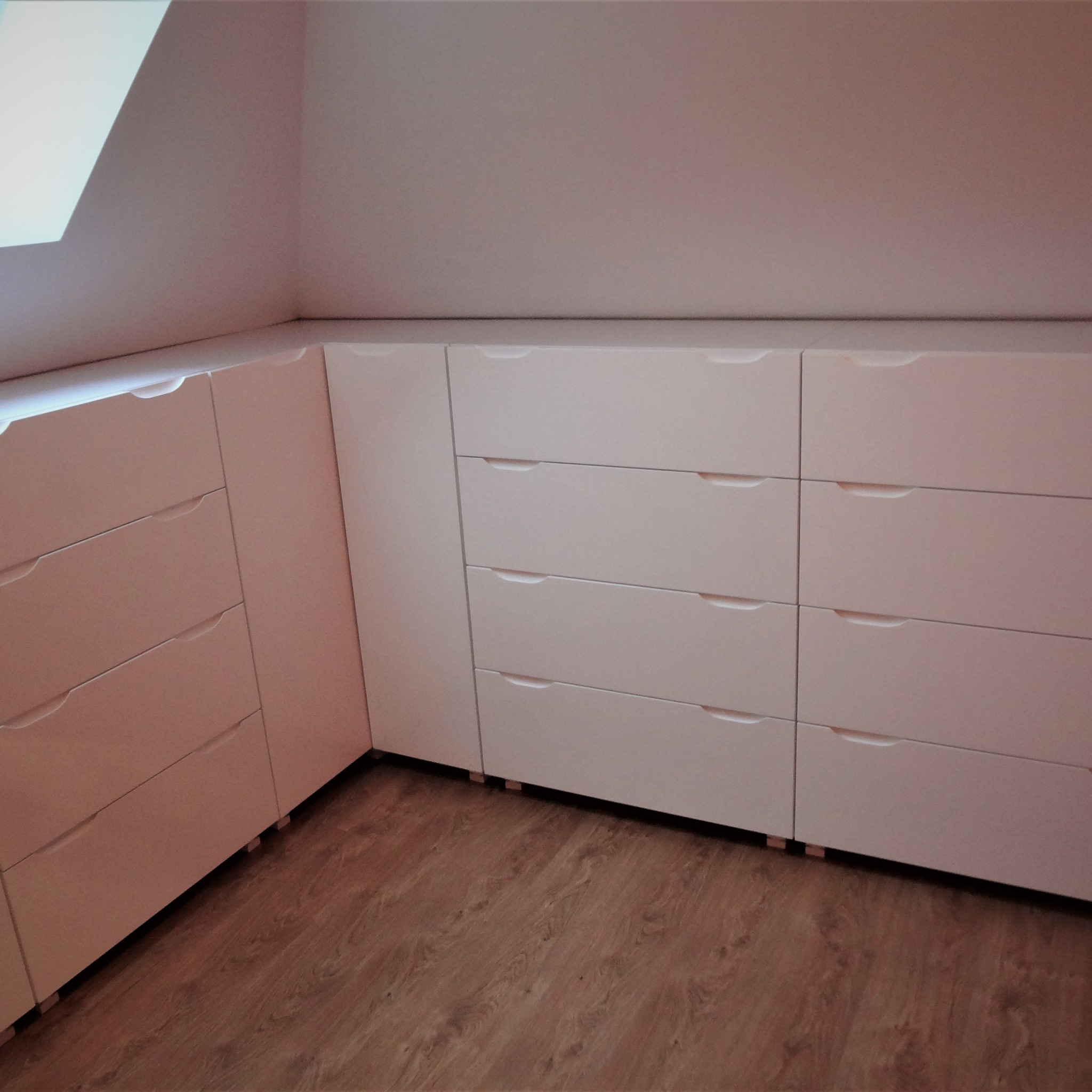 Biała zabudowa pod skosem ze szufladami, wykonana na wymiar w Kwidzynie, z minimalistycznymi uchwytami i drewnianą podłogą.