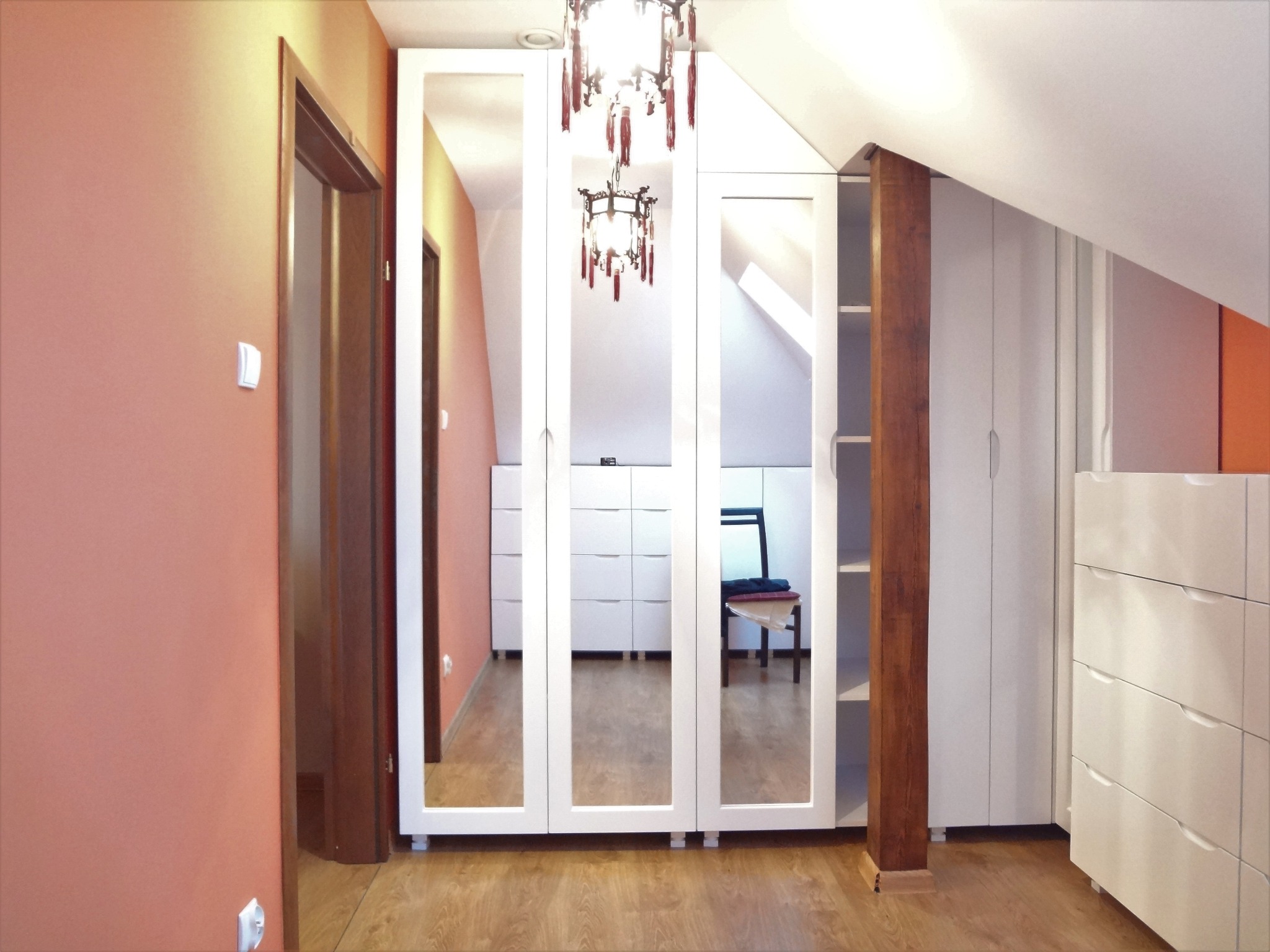 Szafa wnękowa z lustrzanymi drzwiami w skosach poddasza, oświetlona ozdobnym żyrandolem, z widoczną drewnianą kolumną i komodą z szufladami.