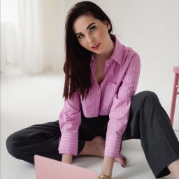 Alicja Szczepańska - Kobieta siedzi na podłodze, ubrana w różową koszulę w paski i szare spodnie, używa różowego laptopa, spoglądając w górę.