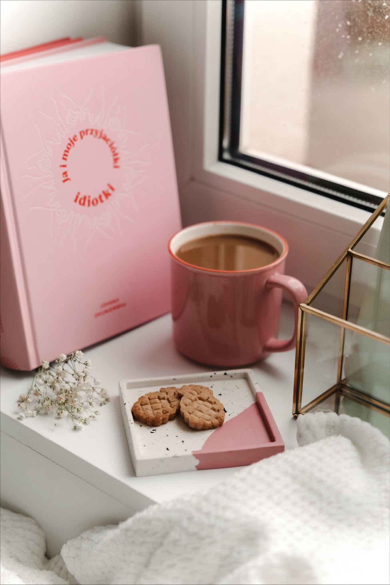 Kompozycja na białym parapecie: różowy kubek z kawą, dwa ciasteczka na biało-różowej podstawce, książka w różowej okładce, bukiecik gipsówki i geometryczna szklana ozdoba na tle okna.