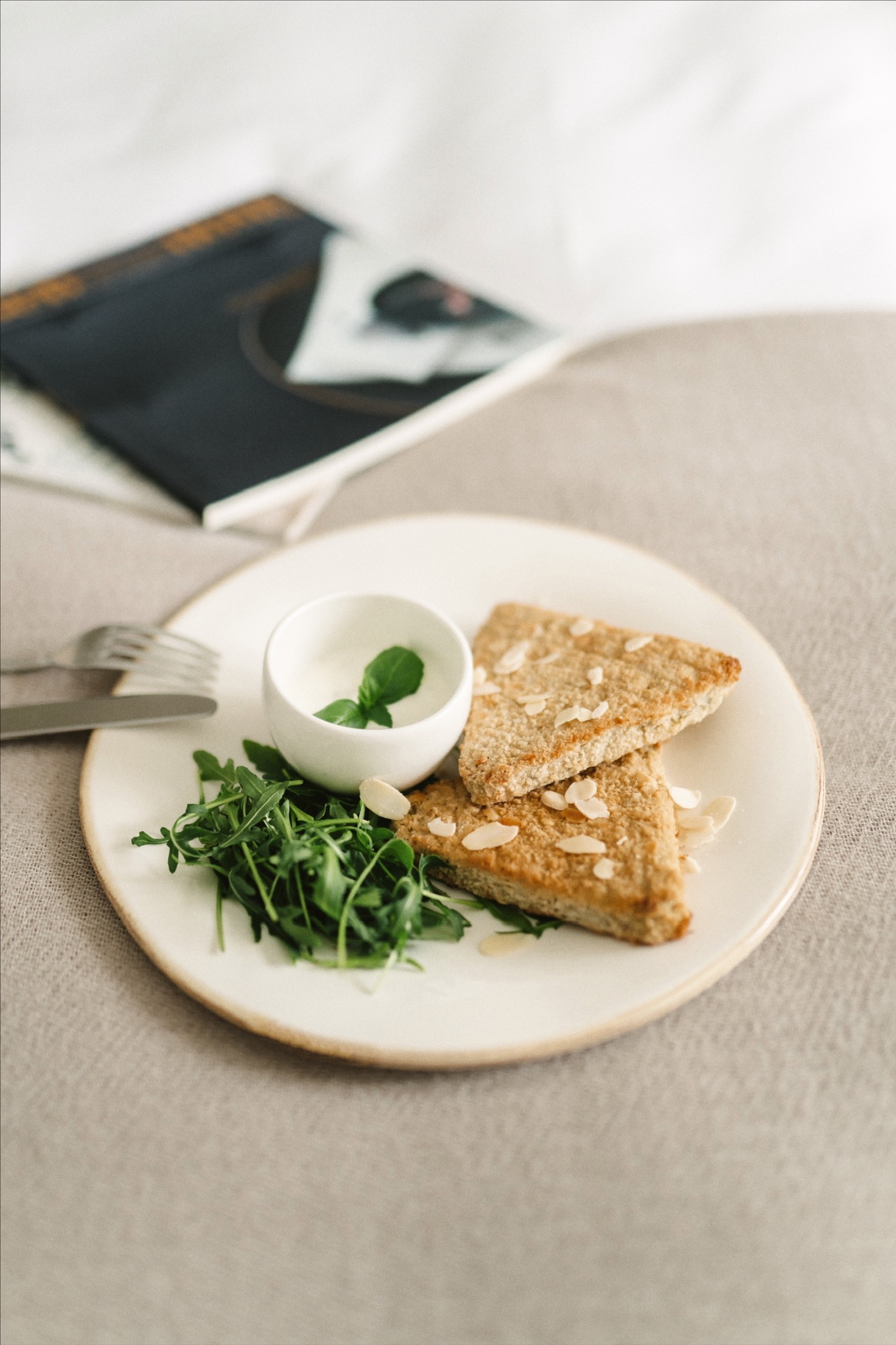 Dwa trójkątne tosty posypane płatkami migdałów, podane na białym talerzu z sosem w małej miseczce i rukolą, obok widelec i nóż, w tle książka.