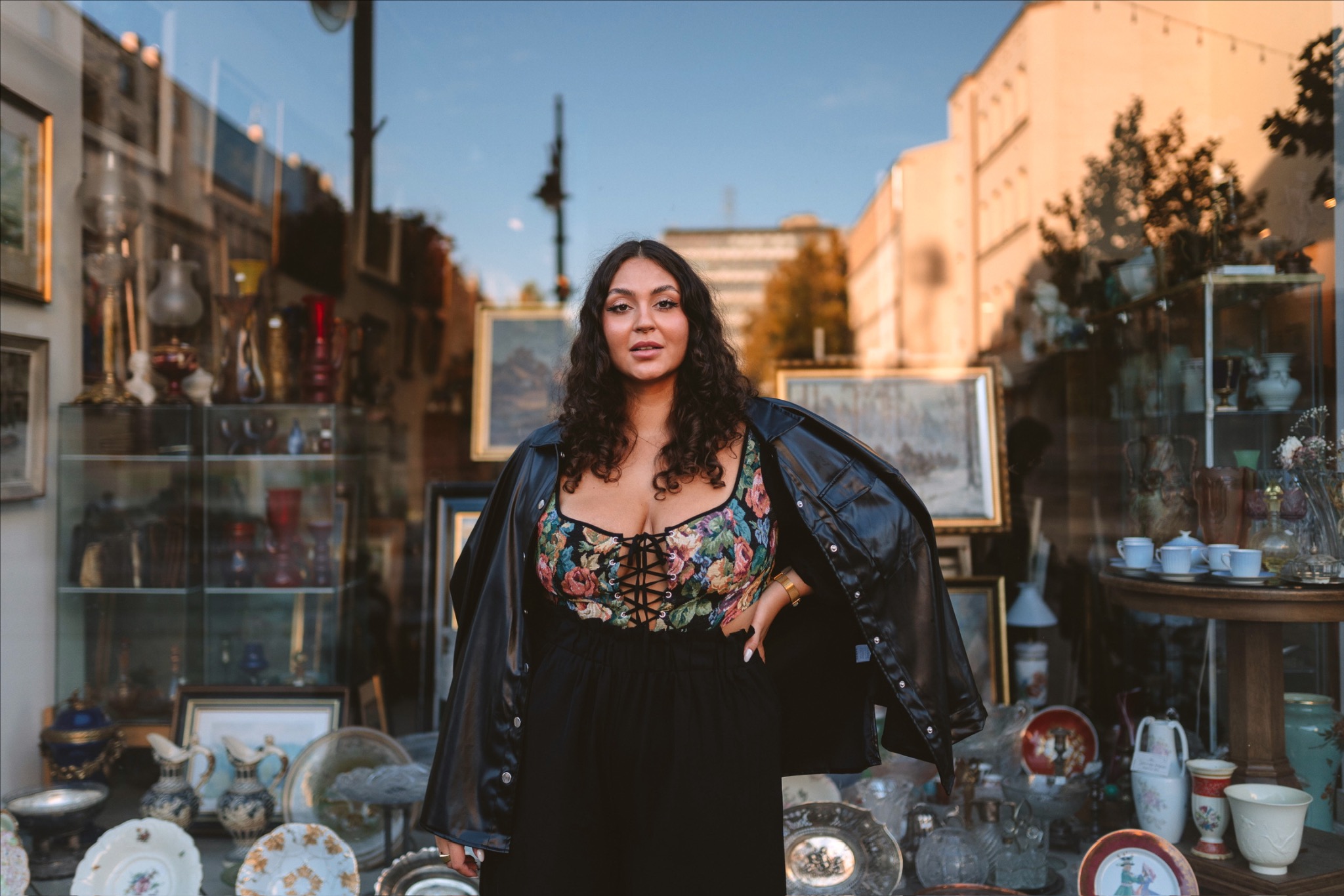 Kobieta w kwiecistym gorsecie i czarnej kurtce pozuje na tle witryny antykwariatu, odbijającej budynki i niebo.