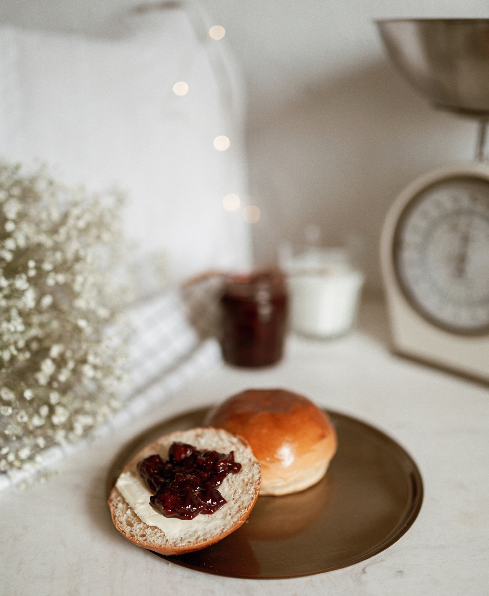 Rozkrojona bułeczka posmarowana masłem i dżemem na metalowej tacy, w tle waga kuchenna, słoik dżemu i gałązka gipsówki, oświetlone delikatnym światłem.