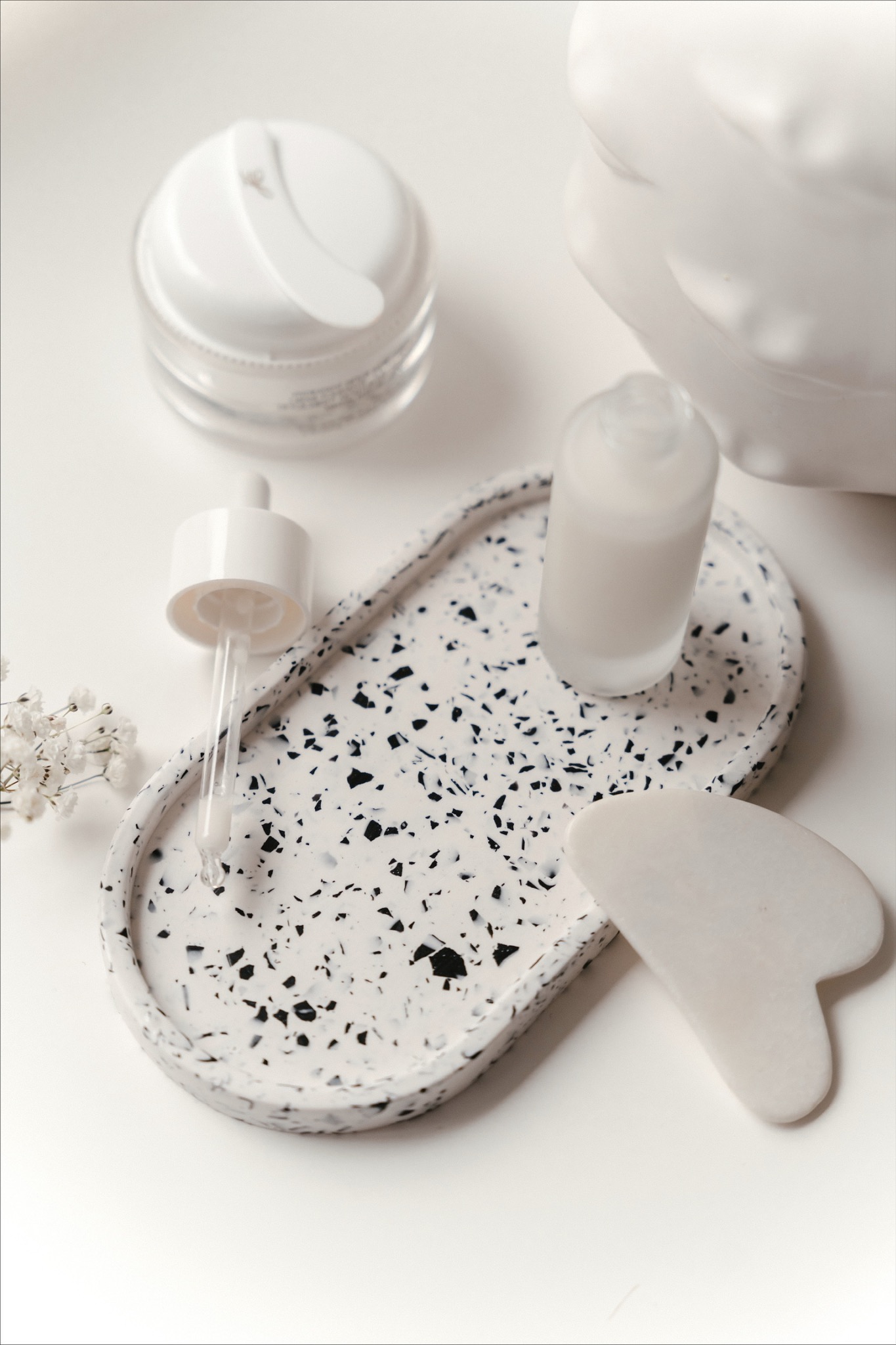 Kompozycja białych kosmetyków na owalnej tacy z wzorem lastryko, obok gipsowa głowa i gałązka gipsówki. Krem w słoiczku, serum z pipetą i kamień gua sha.