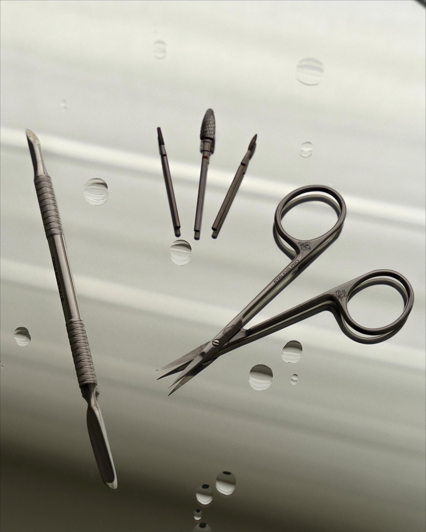 Narzędzia do manicure i pedicure: cążki, frezy, dłutko, ułożone na błyszczącej powierzchni z kroplami wody.