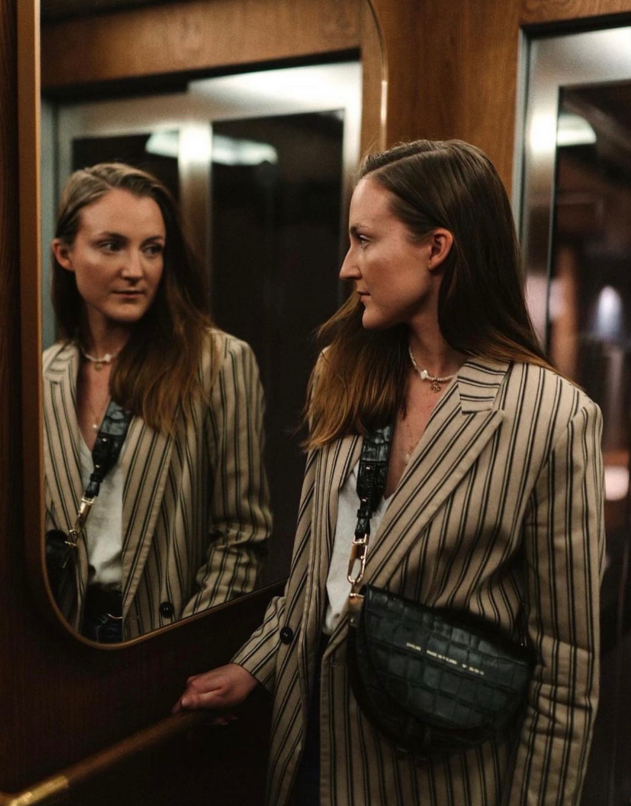 Kobieta w beżowym prążkowanym żakiecie, białej bluzce i czarnej torebce na pasku, pozująca przed lustrem w drewnianej windzie, z delikatnym naszyjnikiem z pereł.