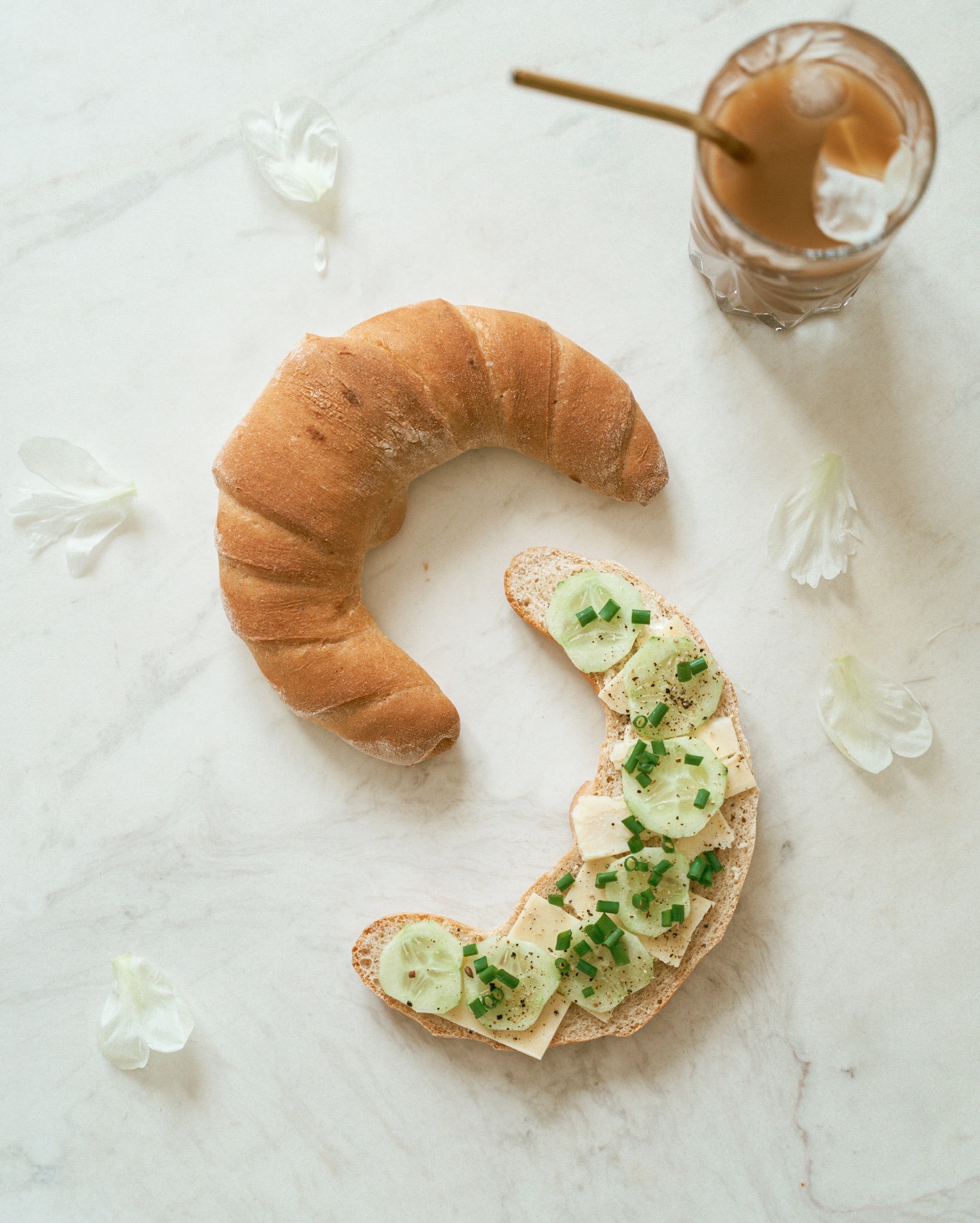 Kompozycja z rogalika, kanapki z serem, ogórkiem i szczypiorkiem oraz kawy mrożonej na marmurowym tle, dopełniona białymi płatkami kwiatów.
