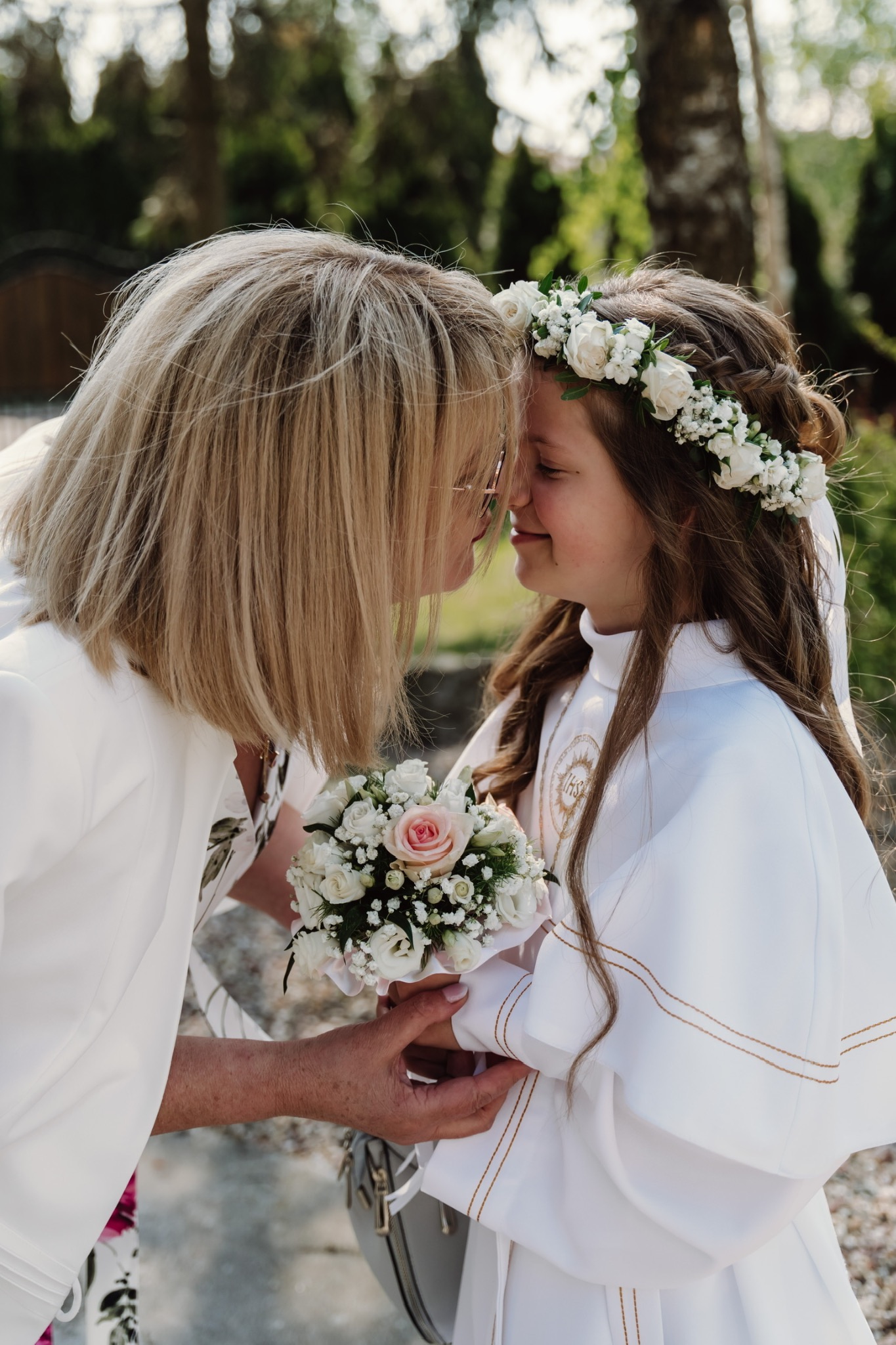 Matka i córka w stroju komunijnym, dotykają się czołami, trzymając bukiet białych róż z jedną różową w centrum, w tle zieleń drzew.