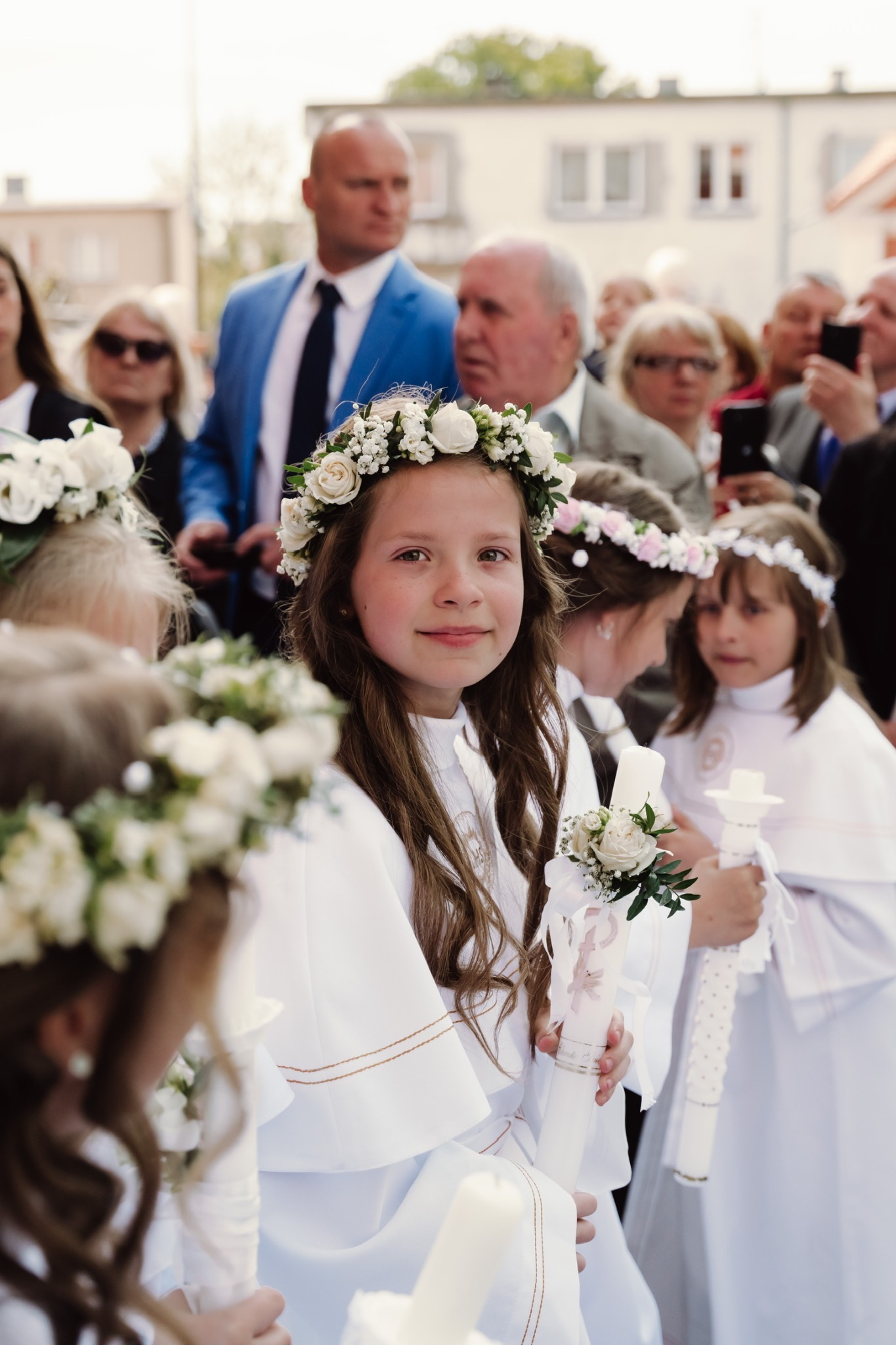 Dziewczynka w białej albie i wianku z białych róż uśmiecha się, trzymając świecę komunijną ozdobioną kwiatami i wstążkami, w tle grupa ludzi.