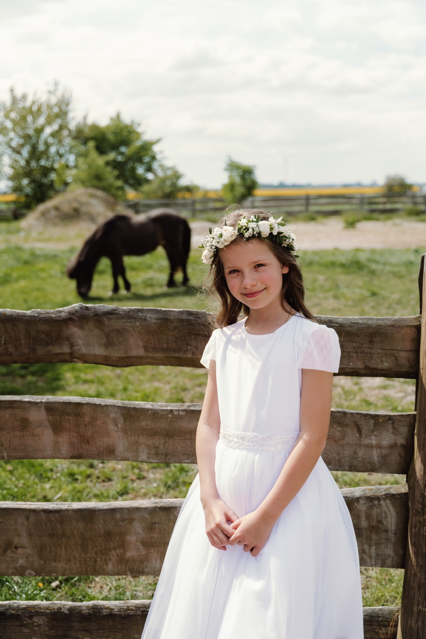 Dziewczynka w białej sukience komunijnej i wianku z białych kwiatów pozuje przy drewnianym płocie. W tle na zielonym pastwisku pasie się ciemny koń.