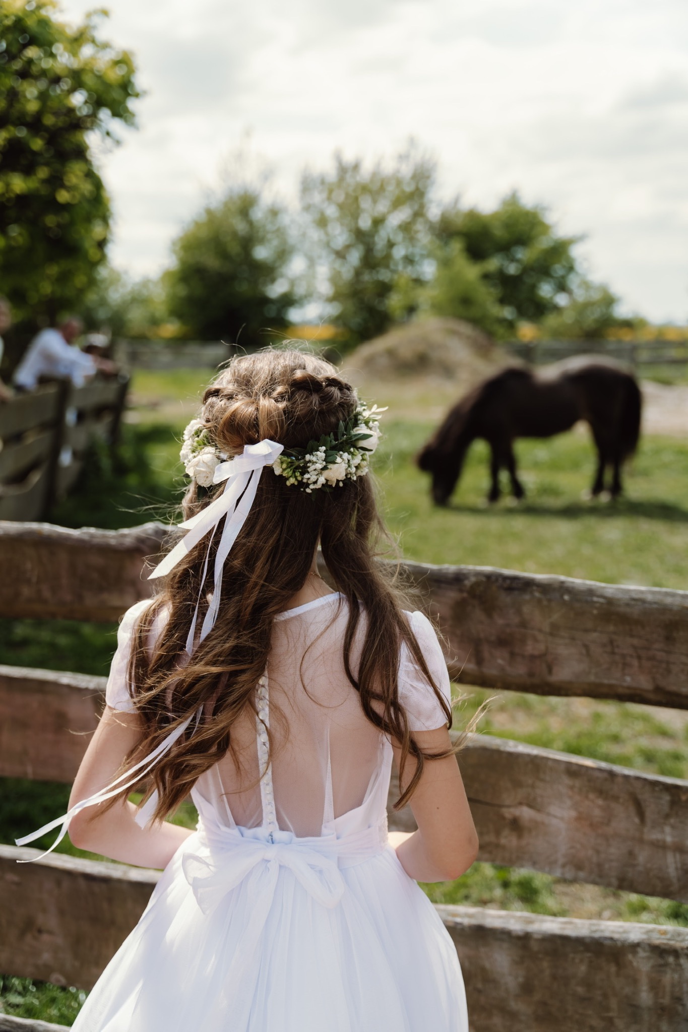Dziewczynka w białej sukience i wianku z białych kwiatów, widziana od tyłu, stoi przed drewnianym płotem, za którym widać pastwisko z koniem i osoby w oddali.