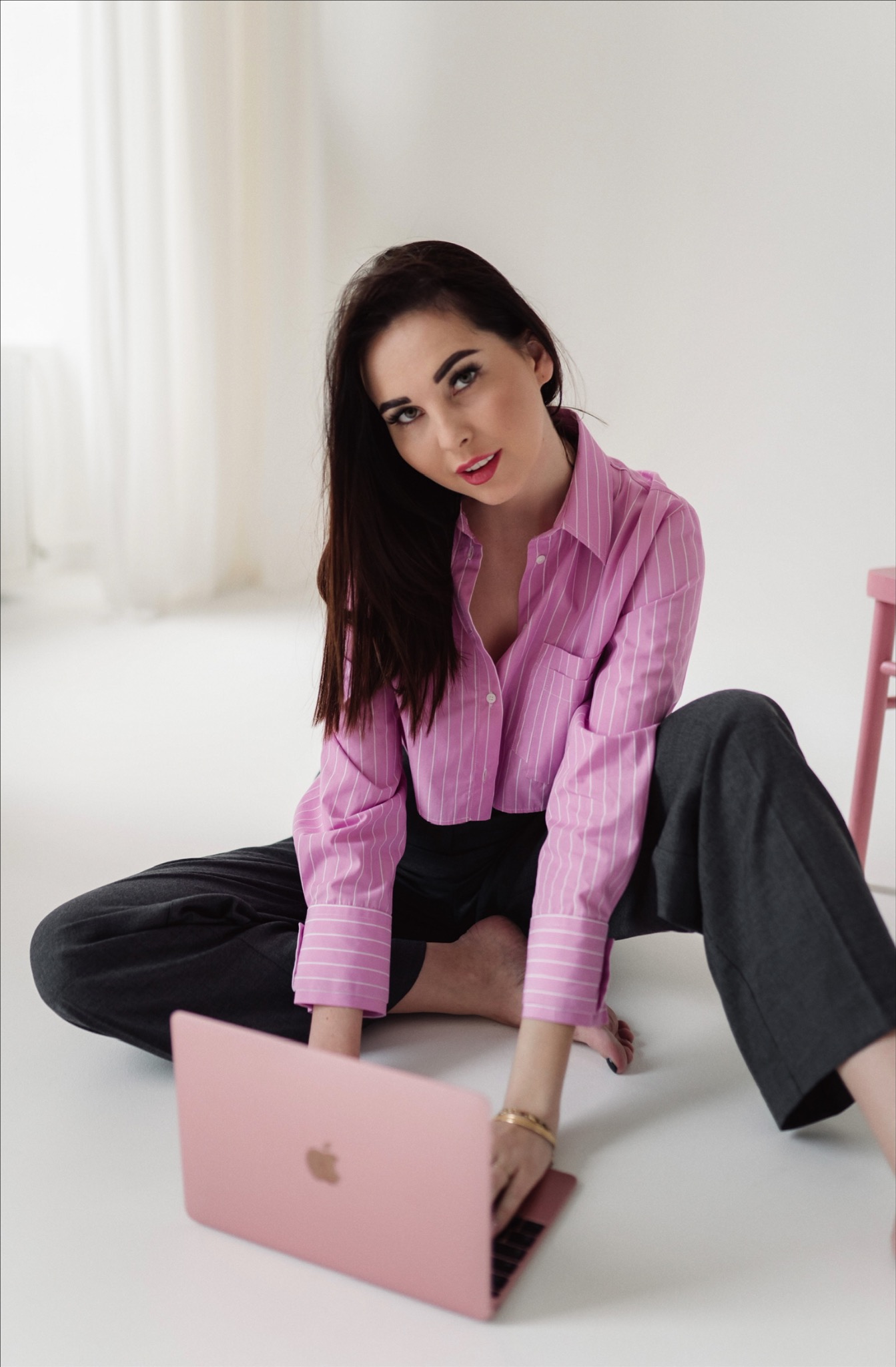 Kobieta siedzi na podłodze, ubrana w różową koszulę w paski i szare spodnie, używa różowego laptopa, spoglądając w górę.