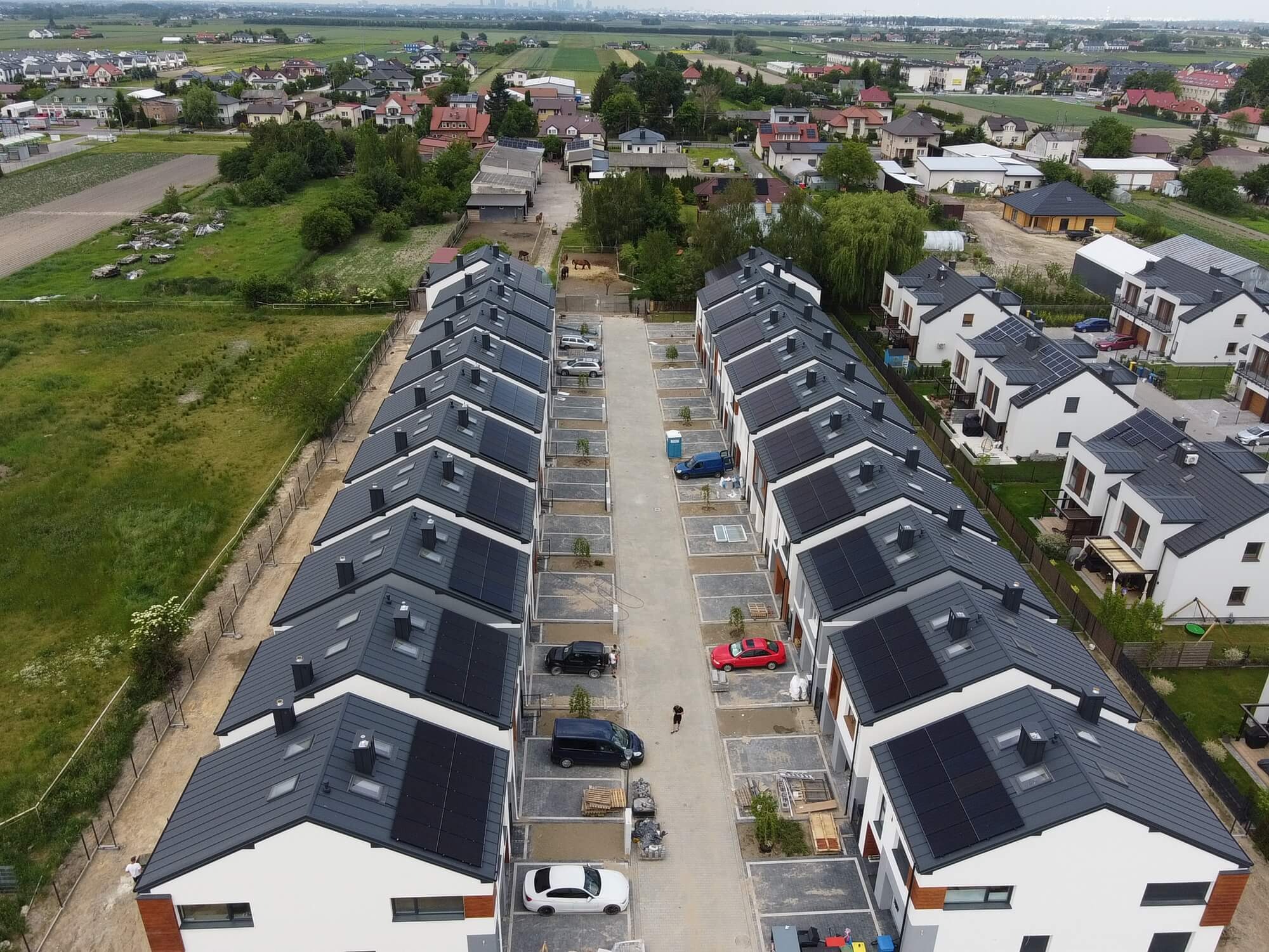 Widok z lotu ptaka na szereg domów jednorodzinnych z panelami słonecznymi na dachach; w tle zabudowa wiejska i odległe miasto.