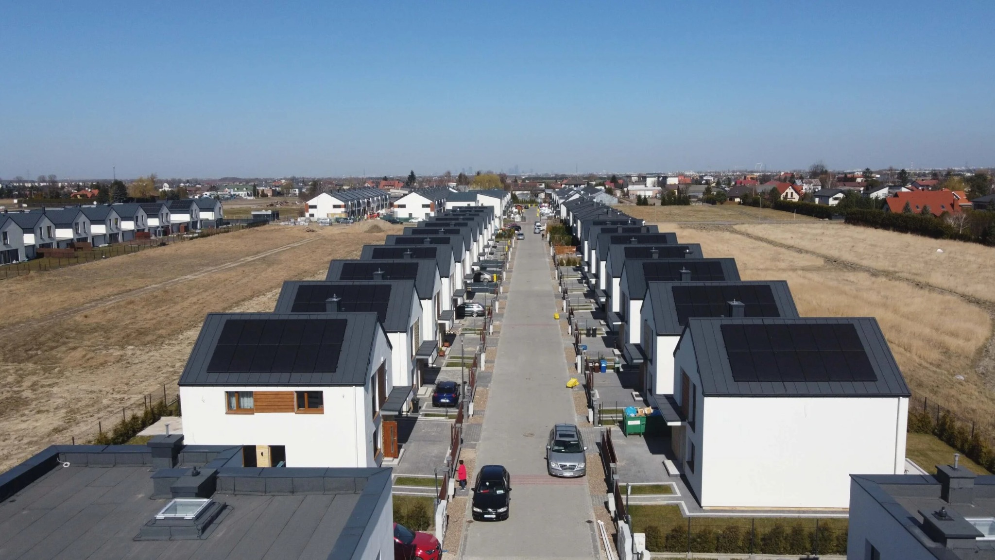 Widok z lotu ptaka na szereg identycznych, nowoczesnych domów z panelami fotowoltaicznymi na dachach, ustawionych wzdłuż ulicy osiedlowej w słoneczny dzień.