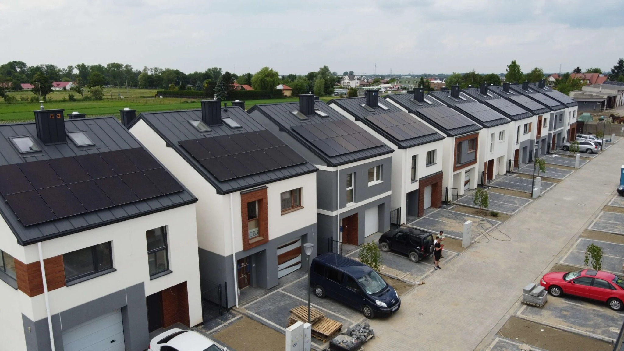 Osiedle nowych domów jednorodzinnych w Łodzi z panelami słonecznymi na dachach, widok z góry, częściowo wykończony chodnik, zaparkowane samochody.