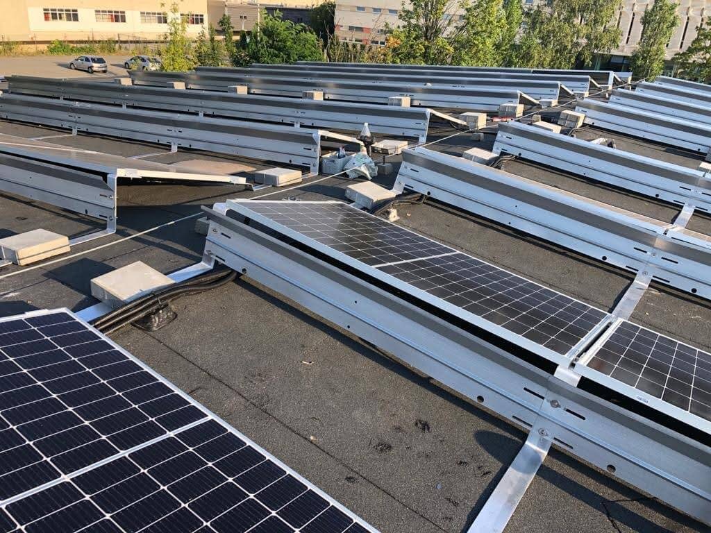 Montaż paneli słonecznych na płaskim dachu, widoczne metalowe profile montażowe i kable, w tle budynek i drzewa.