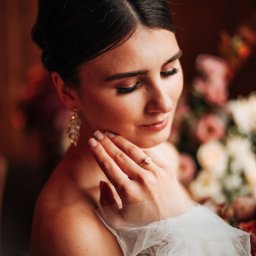Ułamek Miłości - Portret kobiety w sukni z tiulu, z pierścionkiem zaręczynowym na palcu, delikatnie dotykającej szyi na tle bukietu kwiatów.