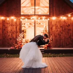Panna młoda w białej tiulowej sukni i sk&oacute;rzanej kurtce, unoszona przez pana młodego w brązowej marynarce, całują się na drewnianym podeście przed rustykalną stodołą ozdobioną girlandami żar&oacute;wek...
