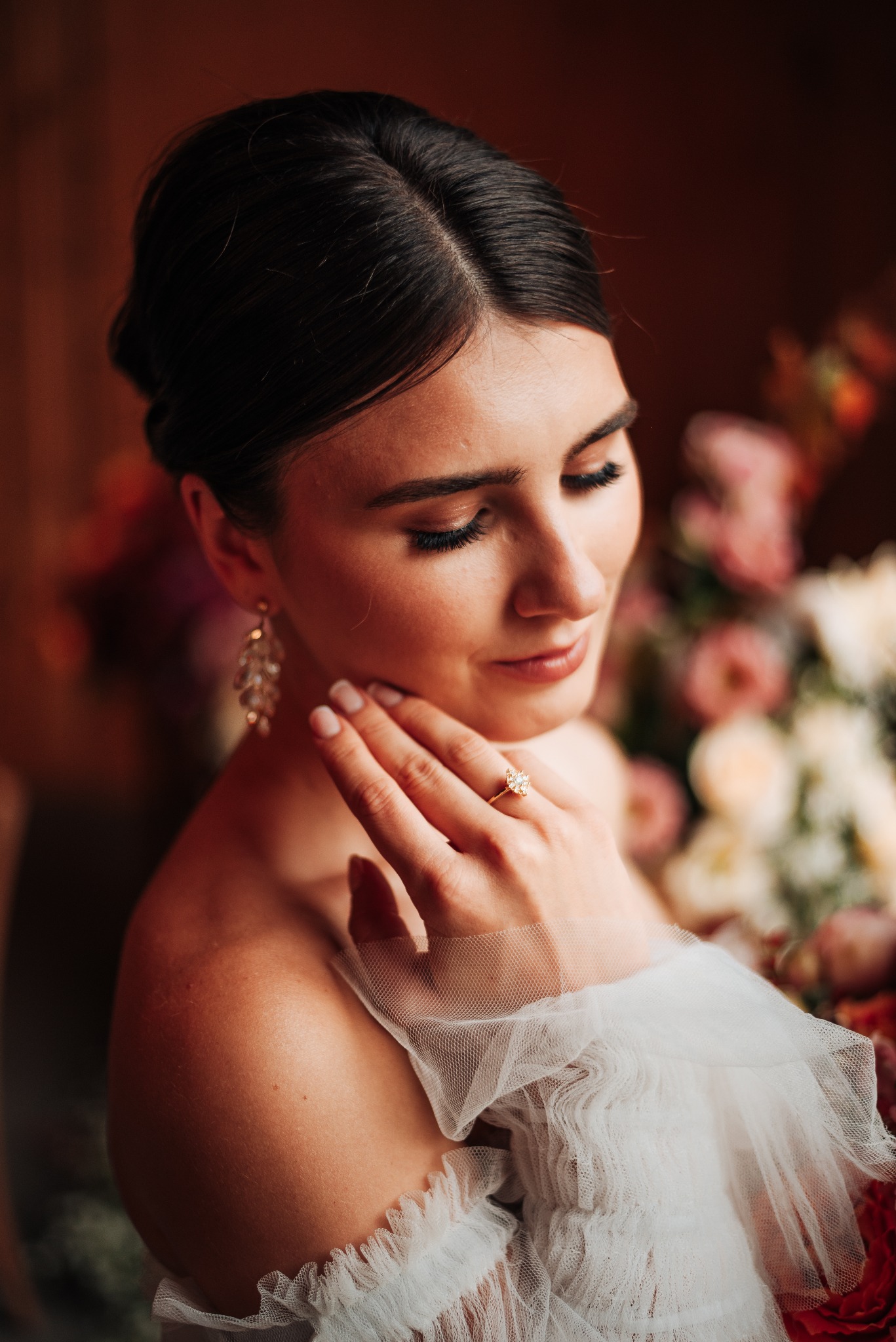 Portret kobiety w sukni z tiulu, z pierścionkiem zaręczynowym na palcu, delikatnie dotykającej szyi na tle bukietu kwiatów.