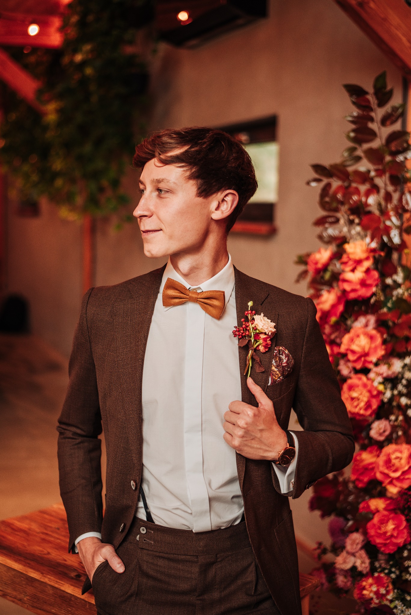 Elegancki mężczyzna w brązowym garniturze i muszce, pozujący na tle dekoracji kwiatowej w odcieniach czerwieni i pomarańczy, z widocznym zegarkiem na nadgarstku i butonierką.