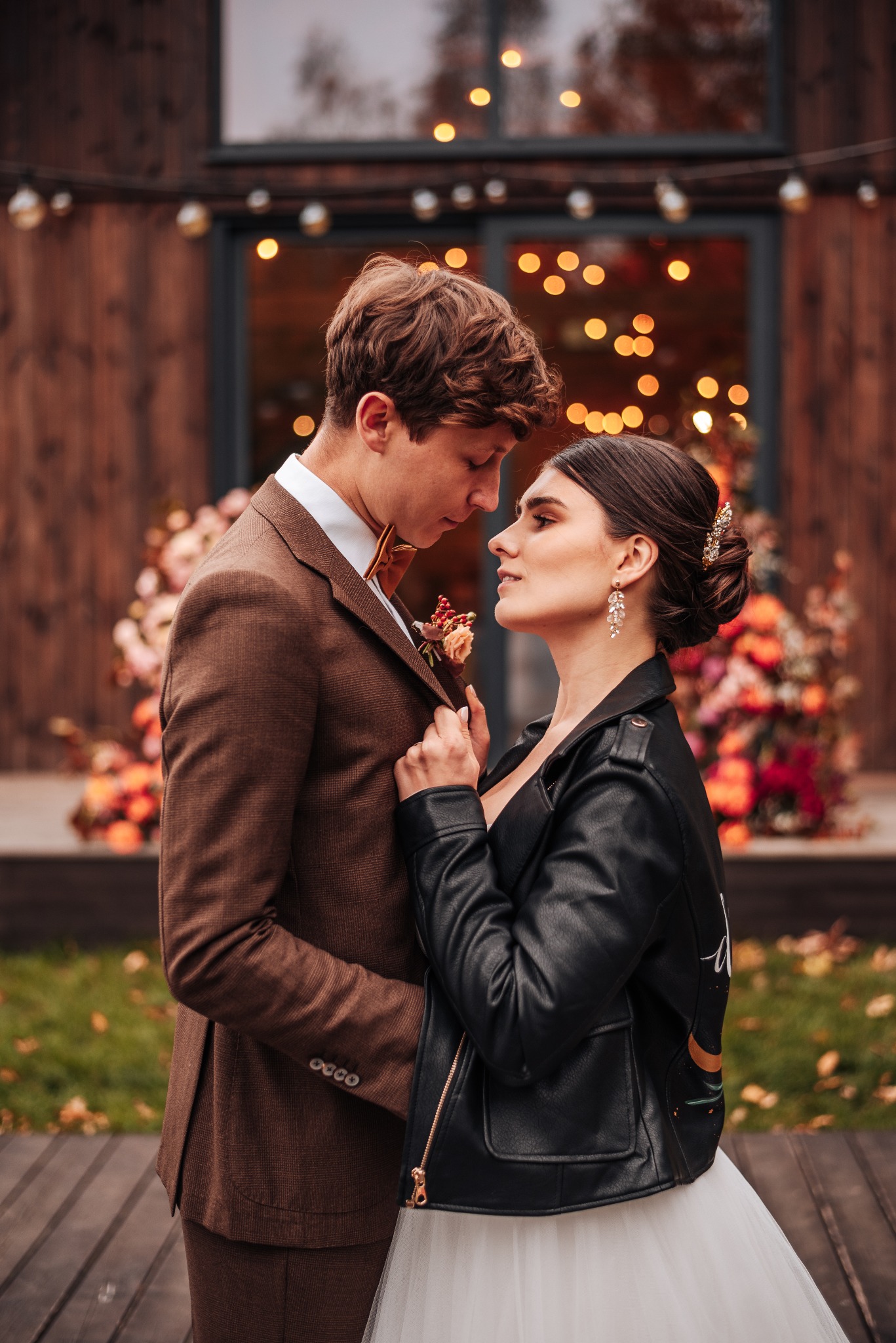 Para młoda w objęciach przed budynkiem z dekoracją kwiatową i światełkami: on w brązowym garniturze, ona w białej sukni i czarnej skórzanej kurtce z malowanymi wzorami na plecach.