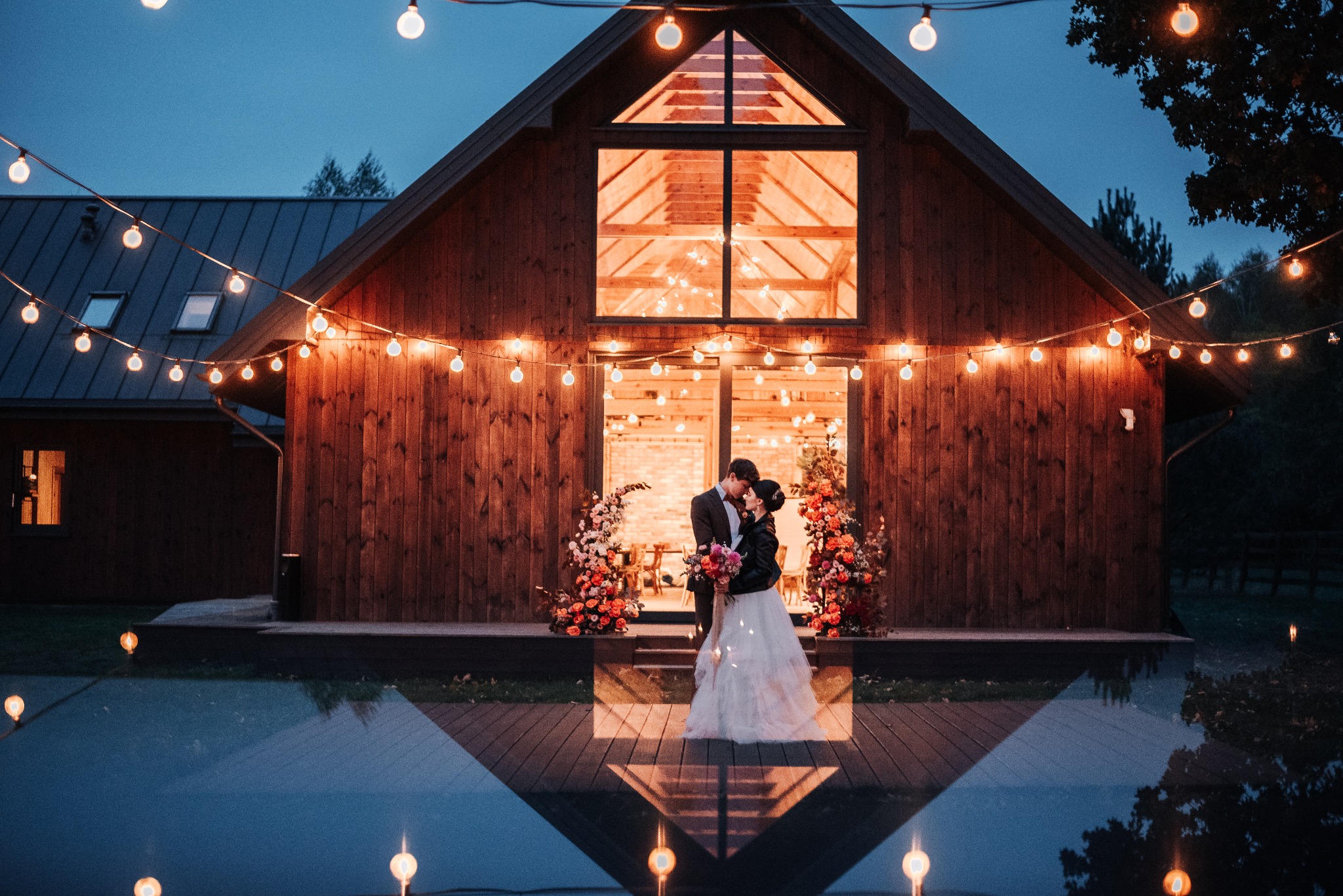 Para młoda całuje się na tle drewnianego budynku z oknem w kształcie trójkąta, udekorowanego girlandami żarówek i kompozycjami kwiatowymi, z odbiciem w wodzie.