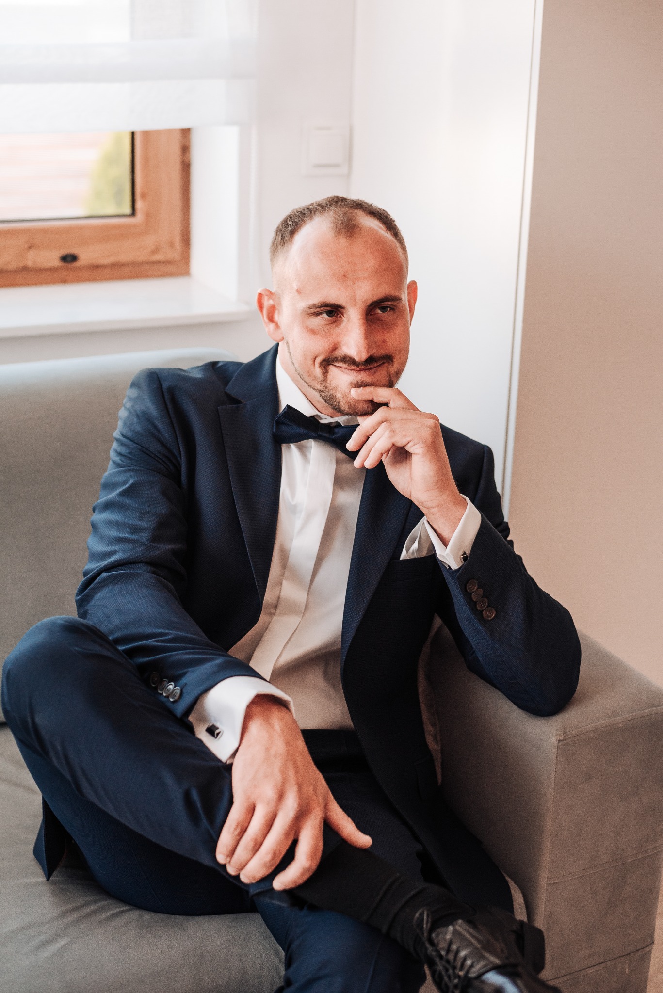 Uśmiechnięty pan młody w granatowym garniturze i muszce, siedzący na szarej sofie podczas przygotowań ślubnych w Bychawie, zamyślony gest dłoni przy brodzie.
