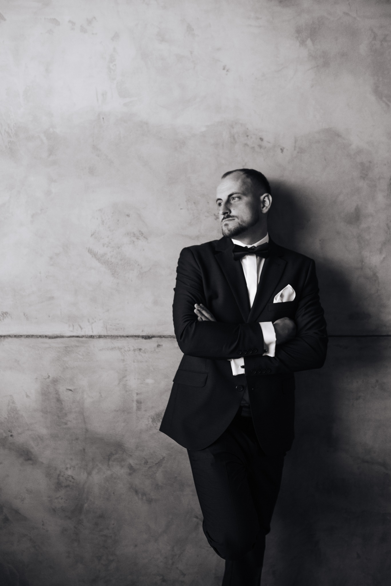 Czarno-białe zdjęcie mężczyzny w czarnym garniturze i muszce, stojącego na tle szarej, teksturowanej ściany z rękami skrzyżowanymi na piersiach i patrzącego w lewo.