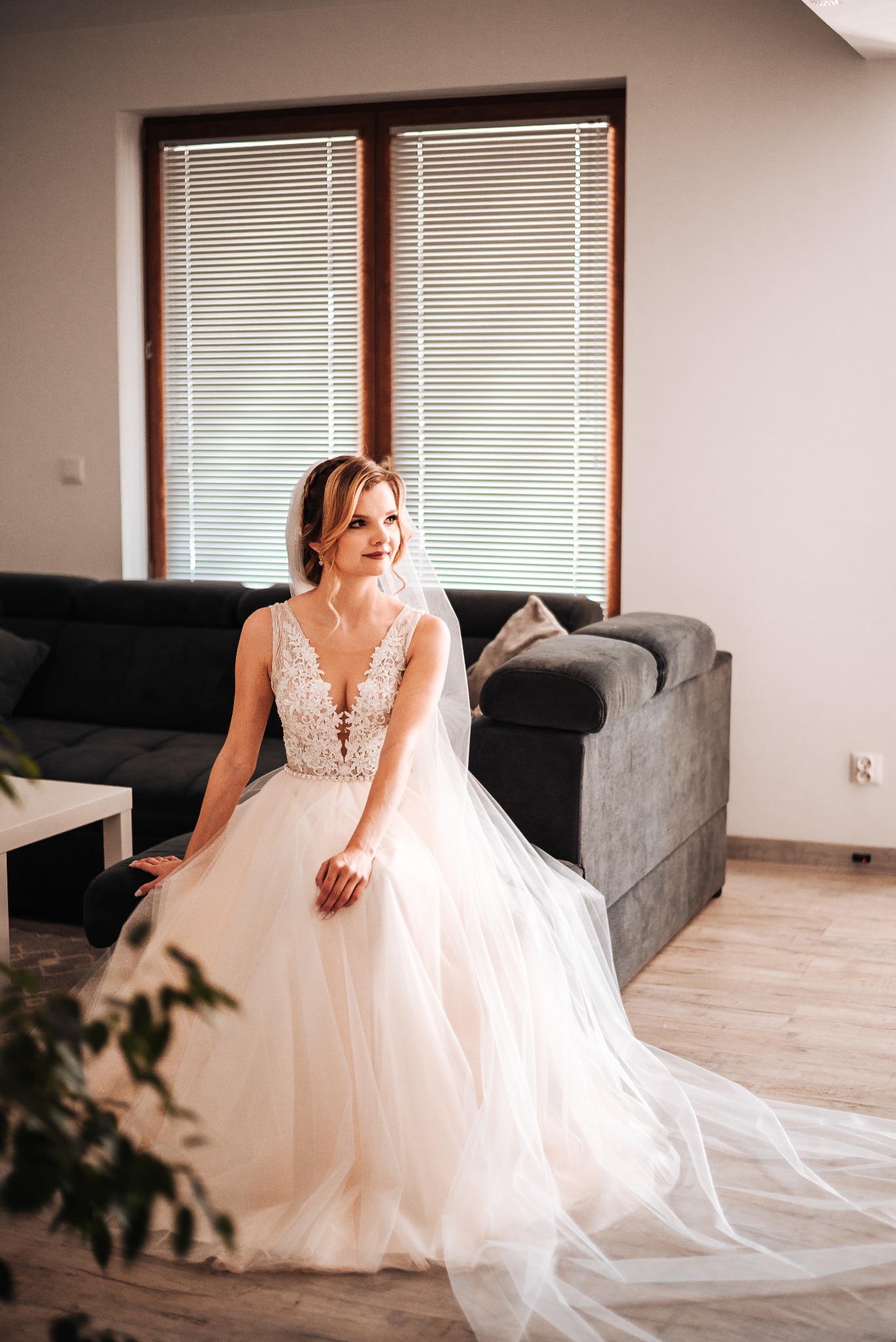 Panna młoda w koronkowej sukni z welonem siedzi na szarej sofie przed oknem z żaluzjami, spoglądając w bok.