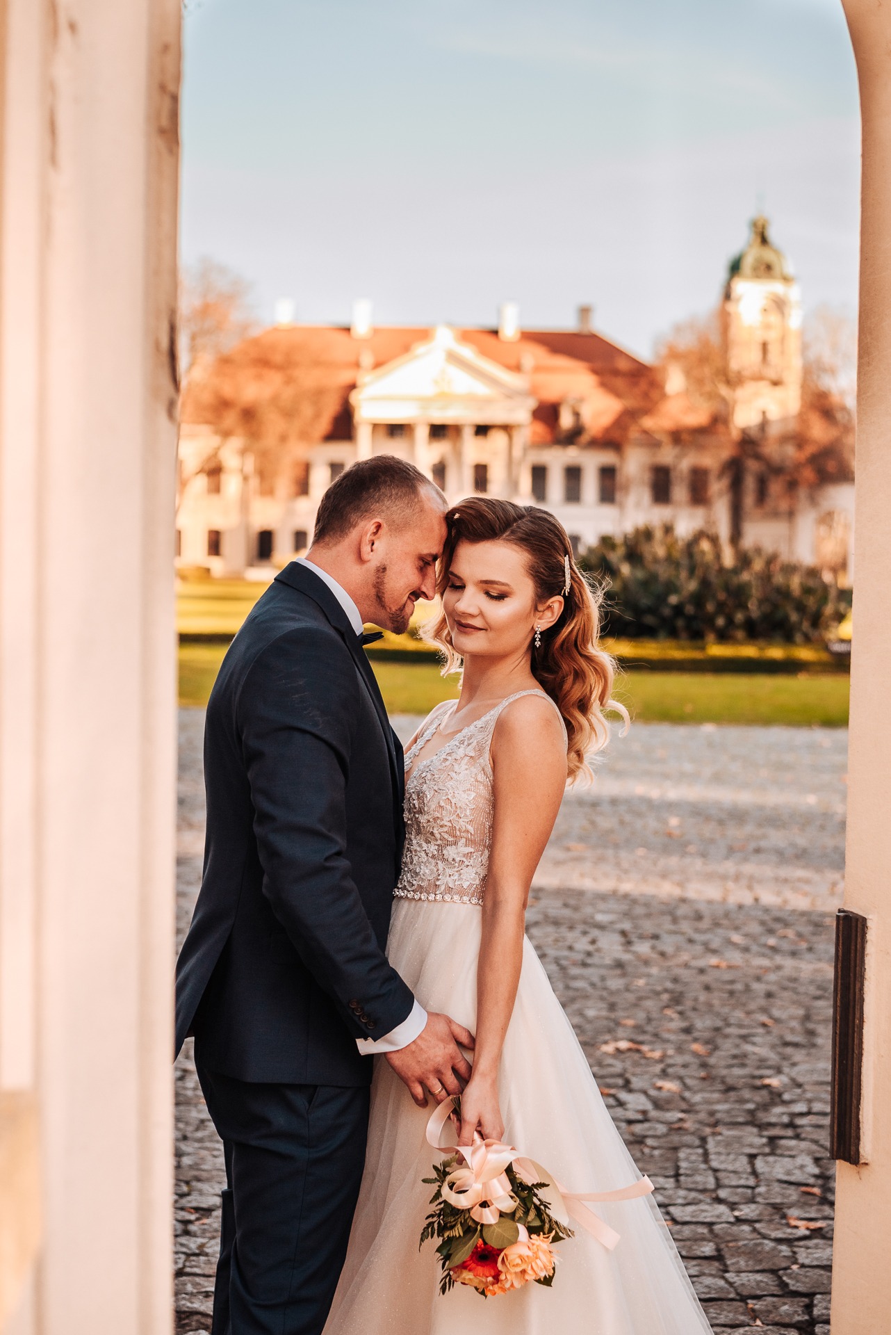 Para młoda, panna młoda w koronkowej sukni z bukietem w pastelowych kolorach, pan młody w granatowym garniturze, stoją przytuleni na tle pałacu z wieżą, widok przez kamienny portal.
