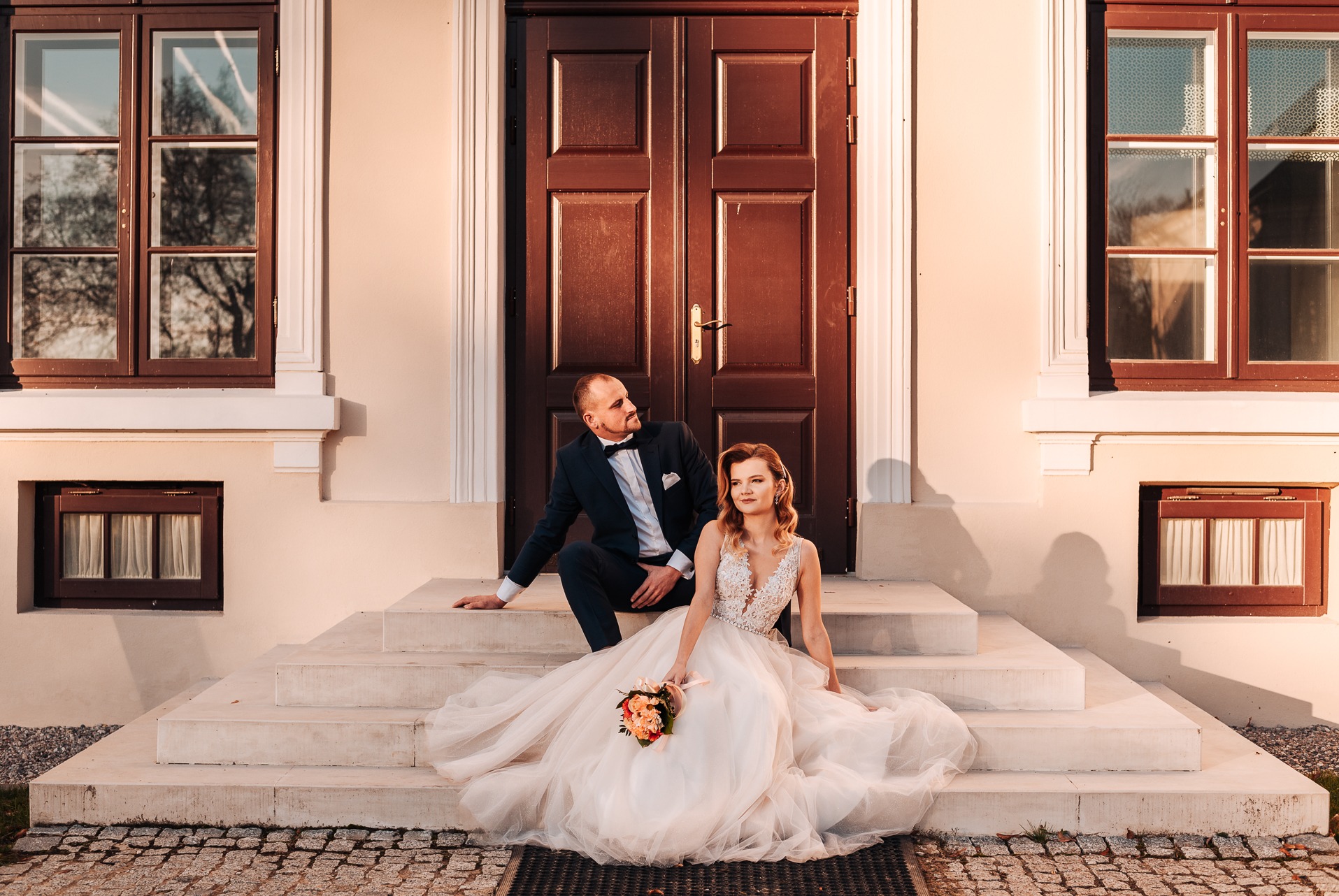 Para młoda siedzi na schodach przed budynkiem z brązowymi oknami i drzwiami. Panna młoda trzyma bukiet, a pan młody patrzy w bok.