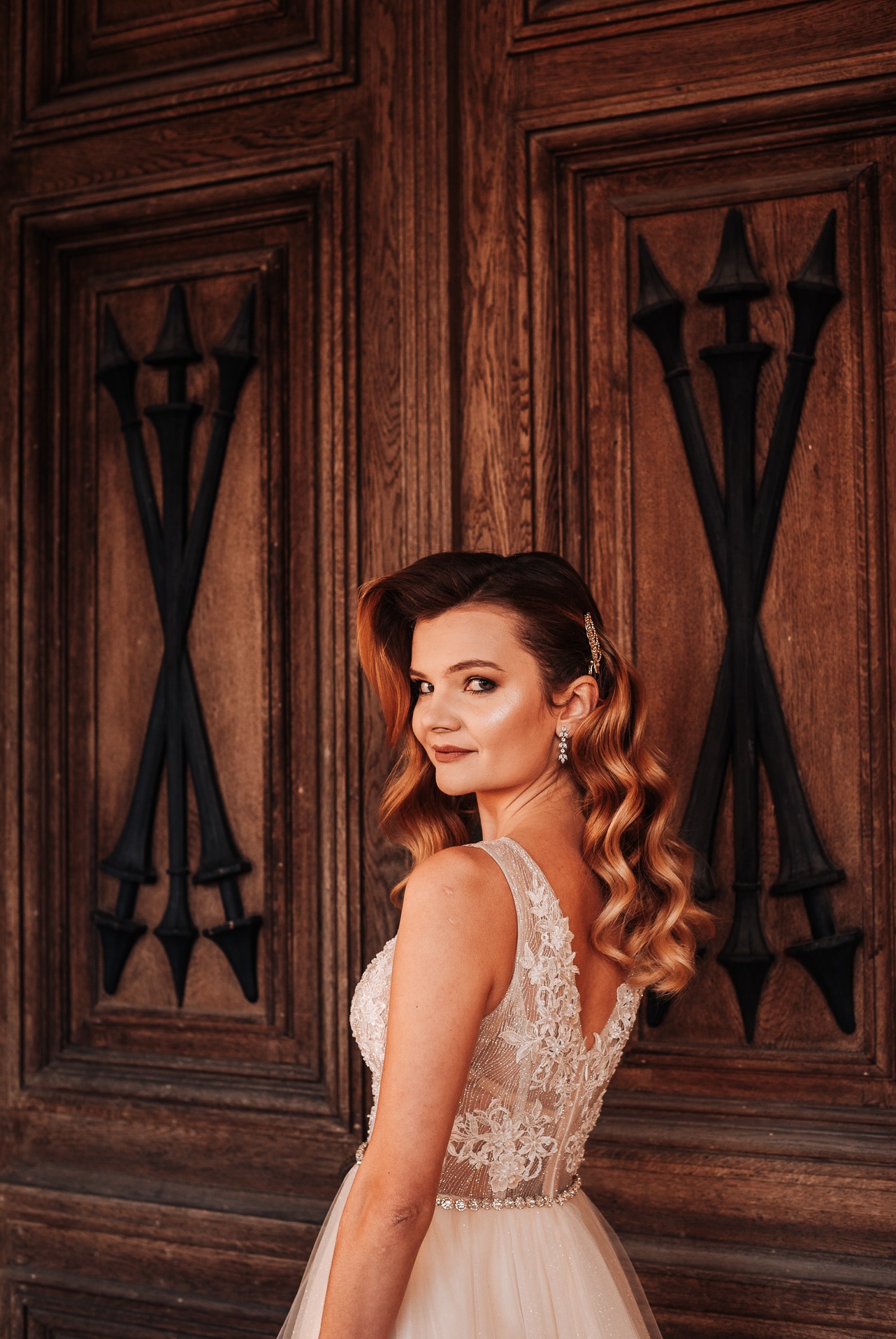 Panna młoda w koronkowej sukni, spoglądająca przez ramię na tle drewnianych drzwi z dekoracyjnymi, czarnymi elementami.